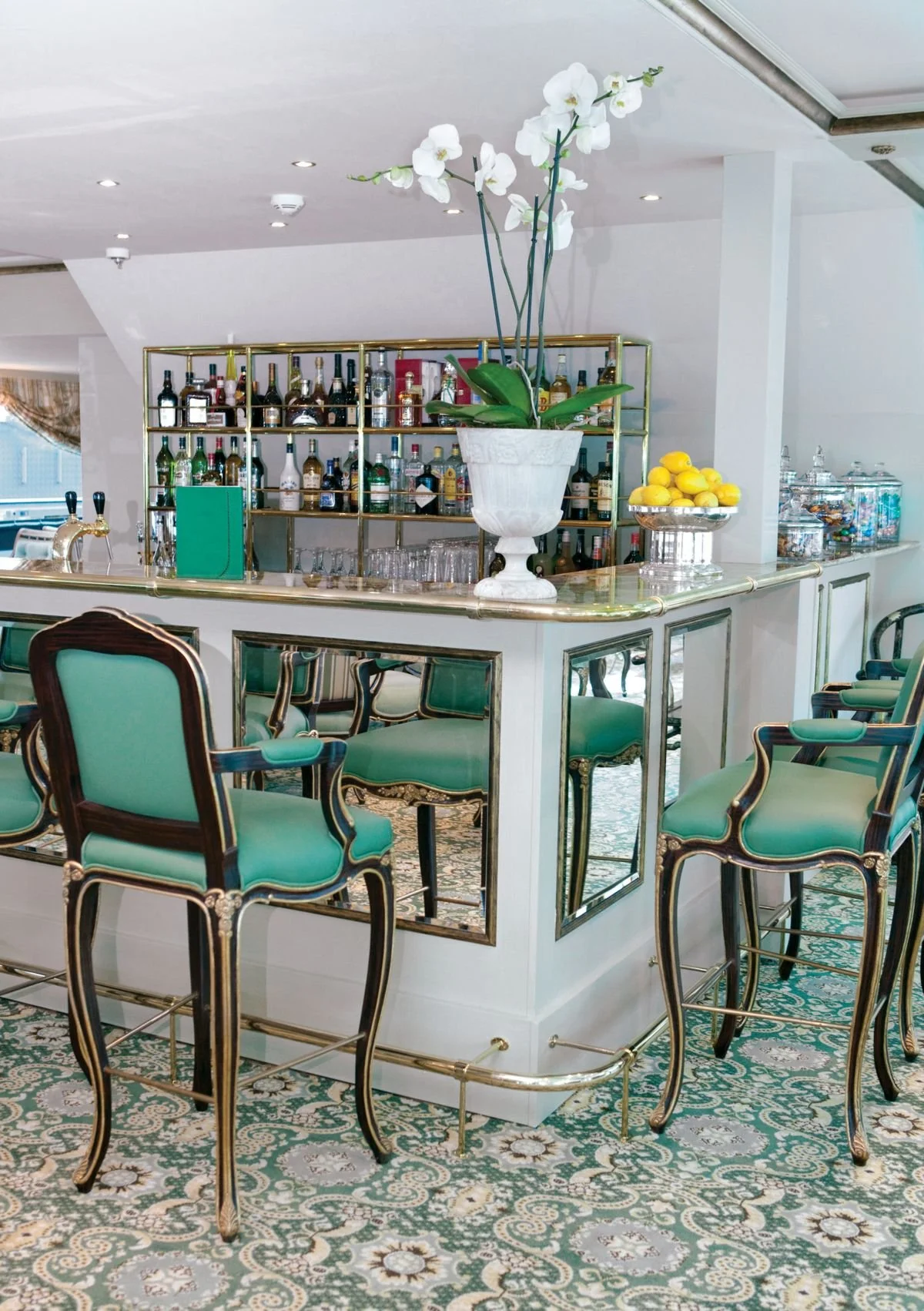A bar area with green upholstered chairs, gold accents, a white orchid in a large white vase, a bowl of lemons, and shelves of liquor bottles.