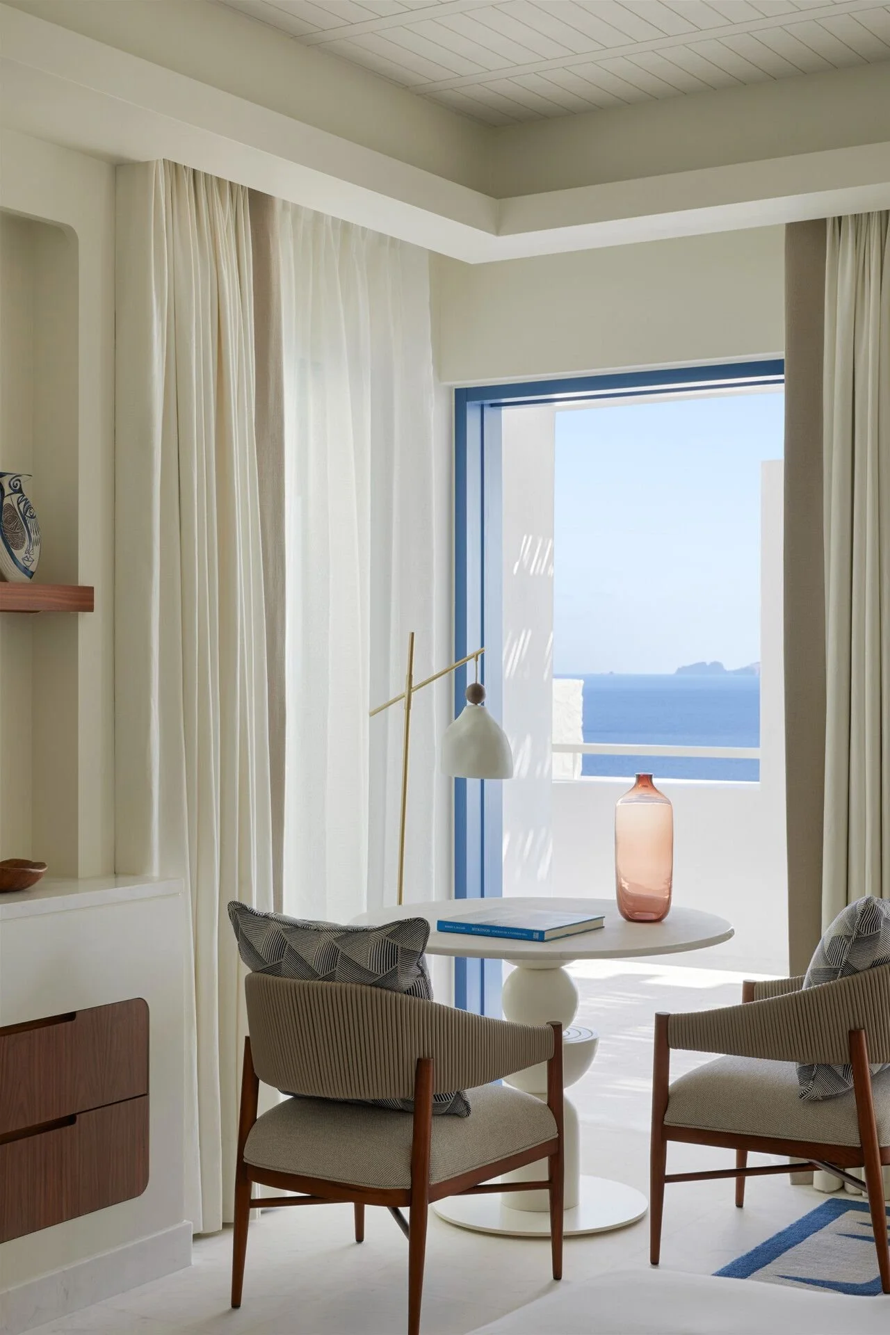 Living room with a round white table, two chairs with patterned cushions, a large window with a view of the ocean, curtains, a pink vase, and a table lamp.