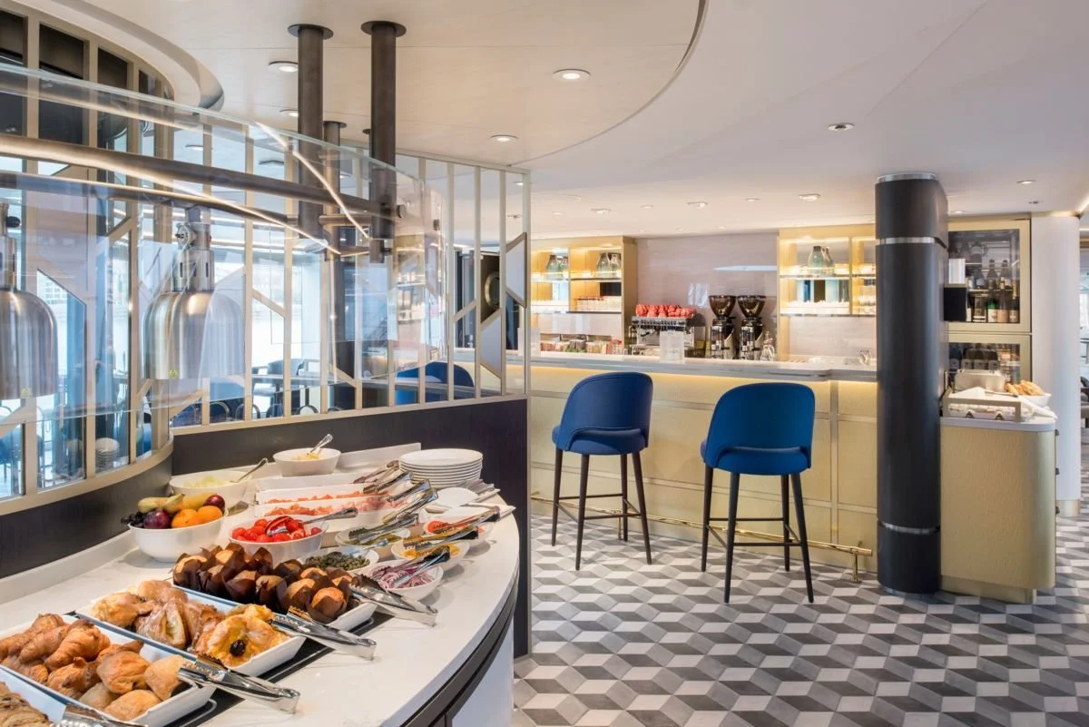 Breakfast buffet with pastries, fruit, and toppings in a modern, well-lit restaurant or hotel lounge.