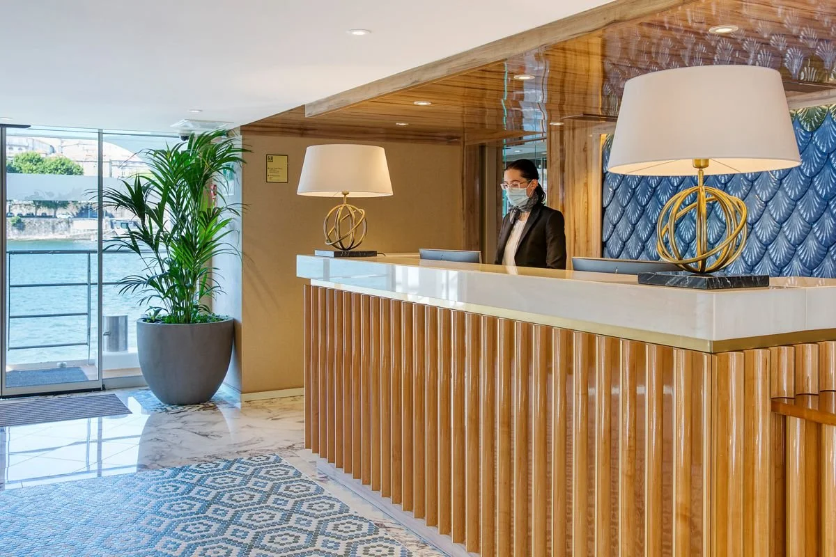 Hotel reception desk with a woman wearing a face mask, black blazer, and glasses standing behind it. There are two large table lamps on the desk, a potted plant by the window, and a decorative blue wall panel. The floor is marble and a geometric-patterned rug is partially visible.