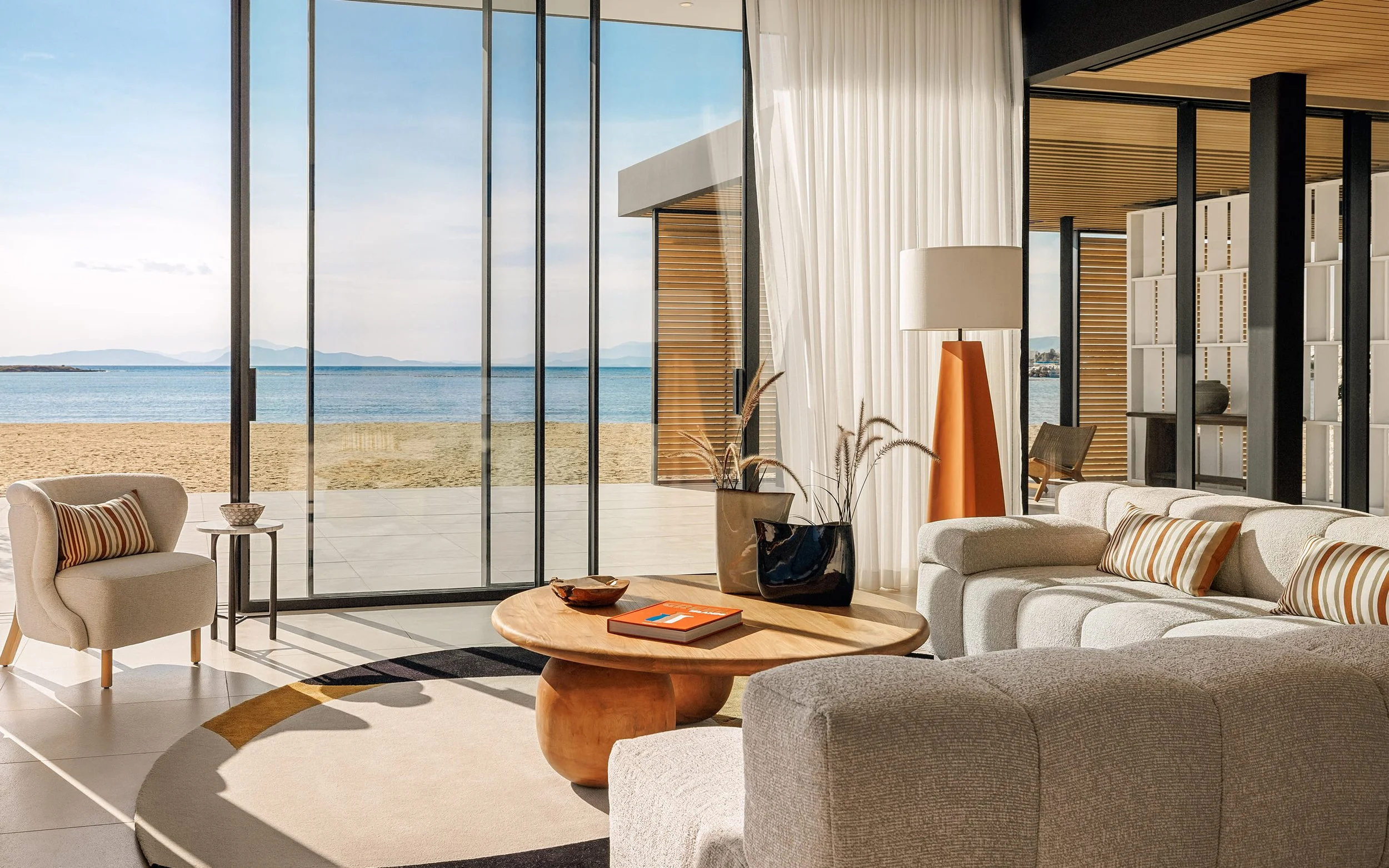 Modern living room with large floor-to-ceiling glass windows showing a beach and ocean view. Furniture includes a beige armchair with a striped pillow, white sofas with striped pillows, a round wooden coffee table with decorative items and books, a tall orange floor lamp, and potted plants. Sheer white curtains hang beside the windows, and the ceiling has wooden paneling.