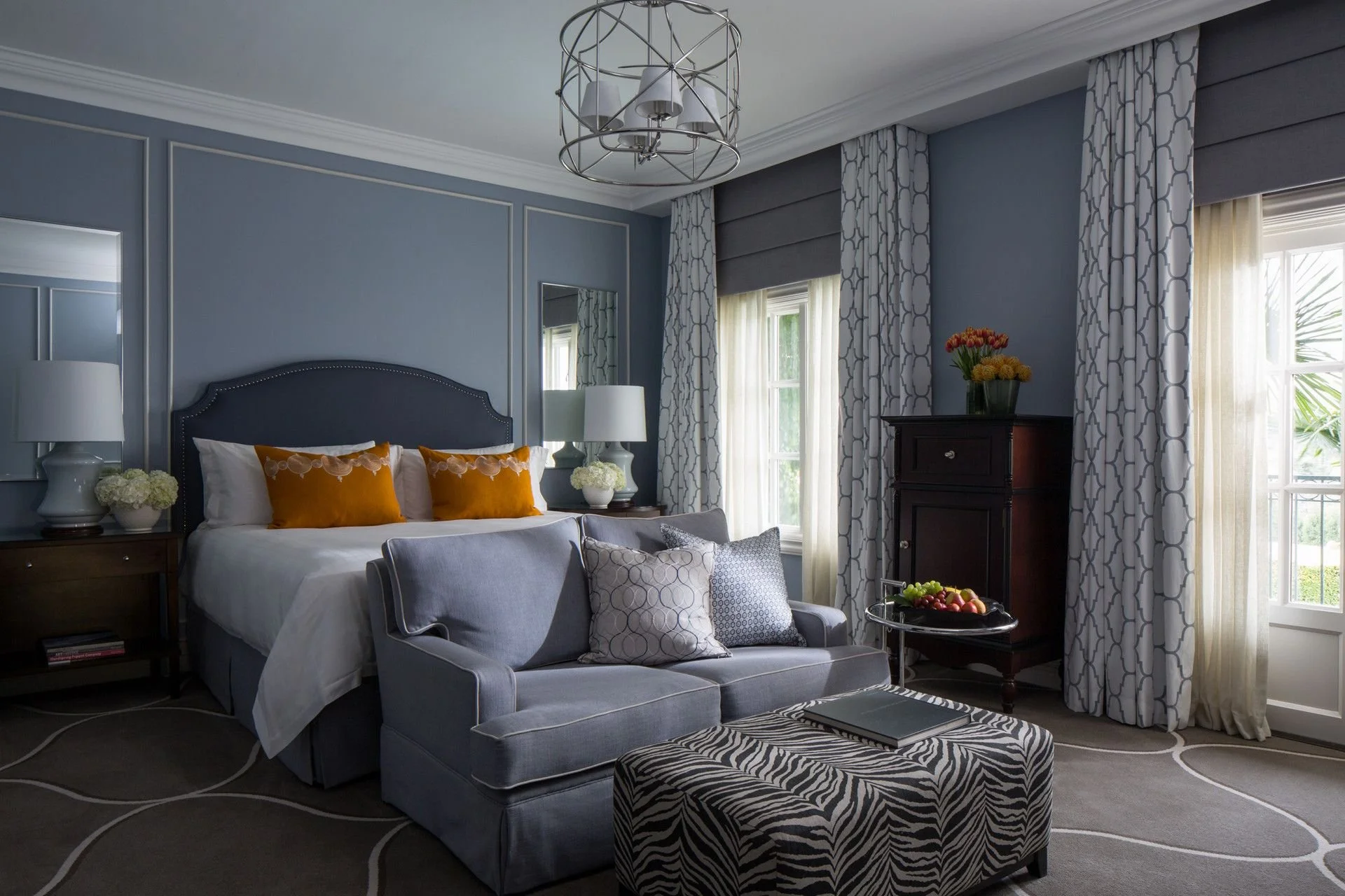 A bedroom with a blue upholstered bed, white bedding, and orange pillows, flanked by two white lamps on nightstands. There's a gray sofa with patterned pillows, a zebra-patterned ottoman, and a black dresser with a vase of flowers. Large windows with patterned curtains allow natural light.