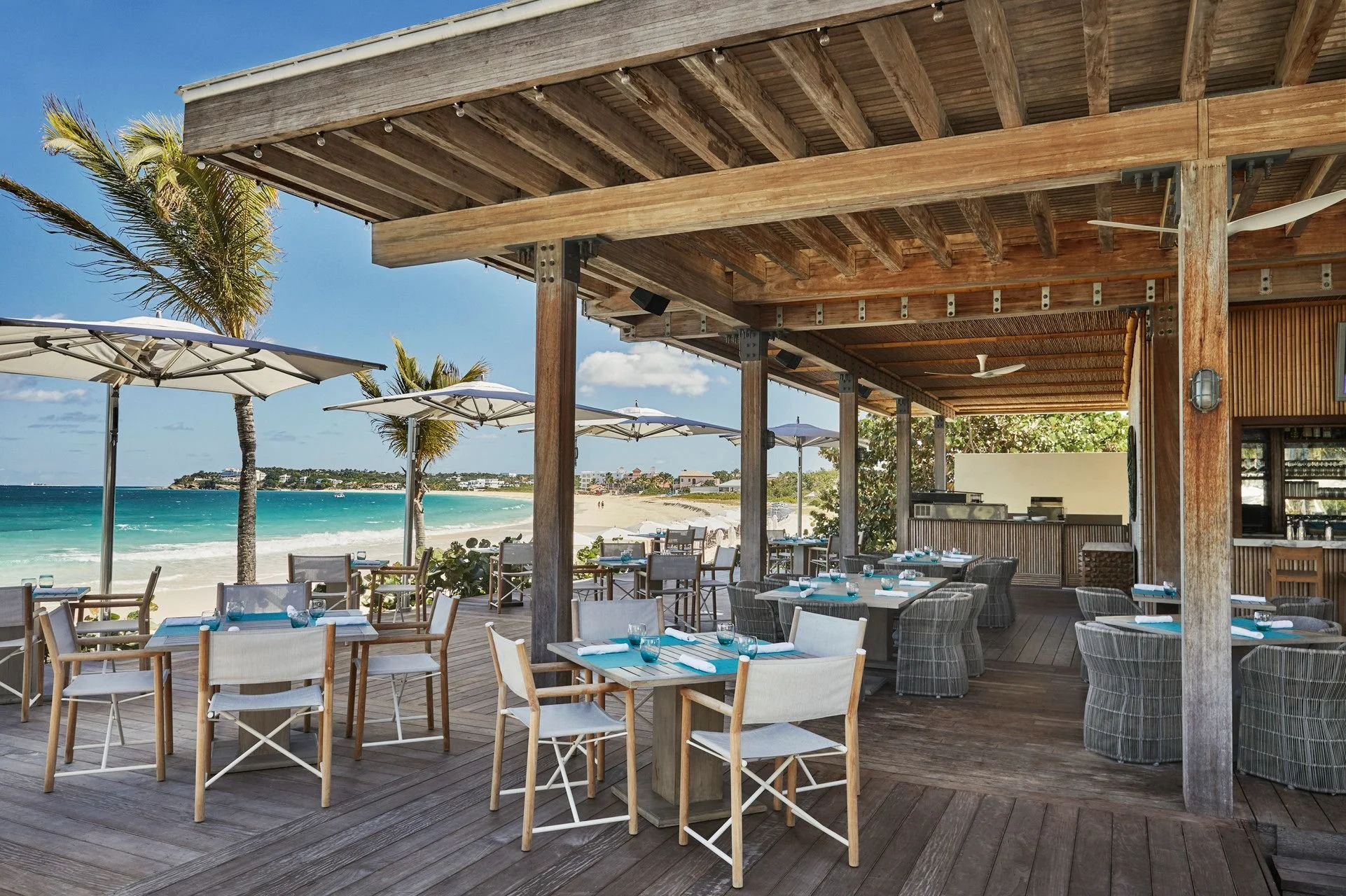 Outdoor restaurant with dining tables set with turquoise glasses and white napkins, located on a wooden deck under a wooden roof. Beach and ocean with umbrellas and palm trees visible in the background.