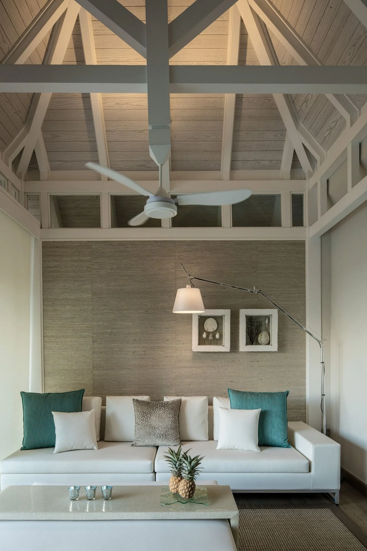 Living room with a white sofa decorated with white and teal pillows, a pineapple centerpiece on a coffee table with three small candles, a beige textured accent wall, a wall lamp, framed decorative items, and a ceiling fan in a room with high wooden ceilings and large windows.