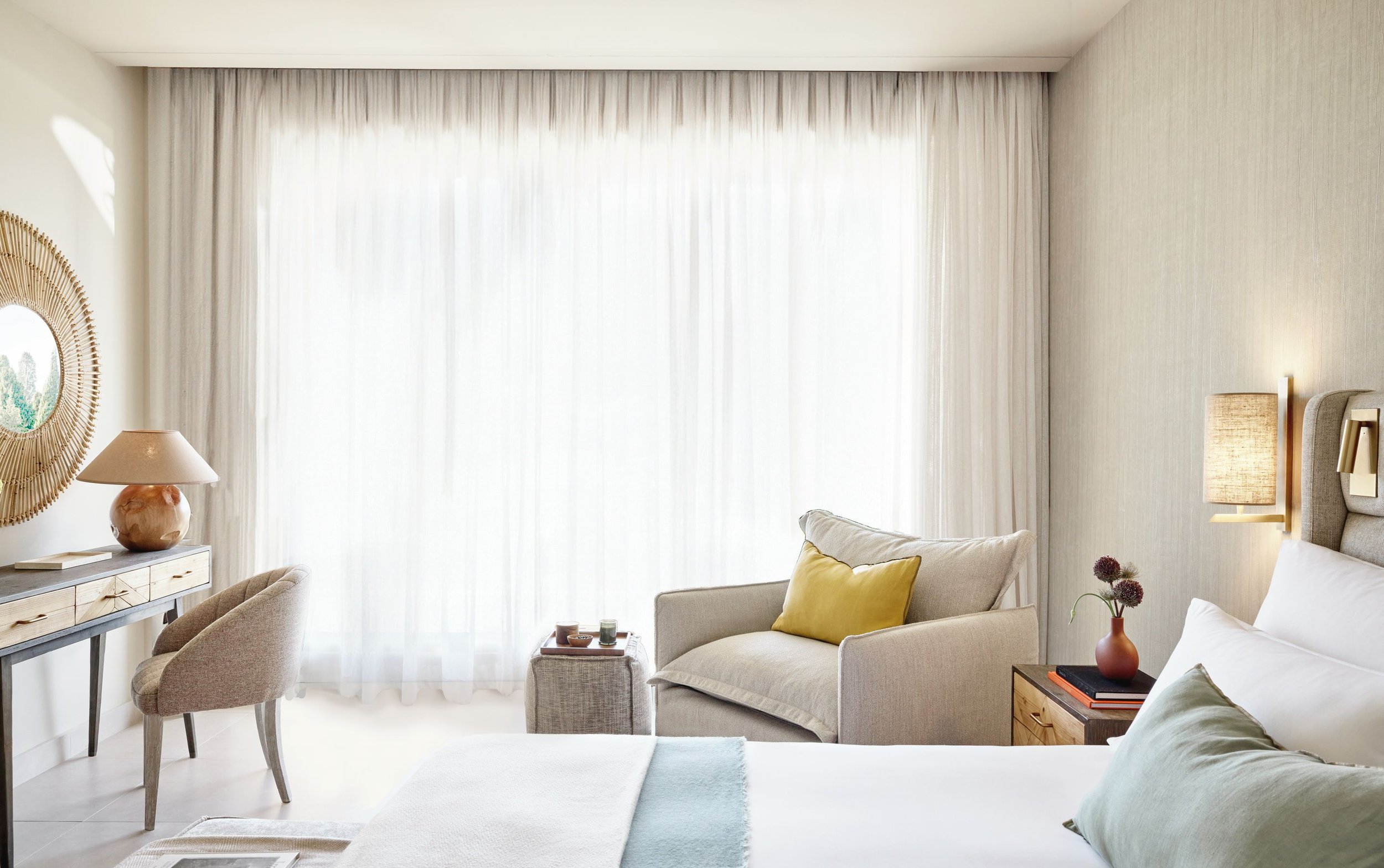 Bright living room with large window covered by sheer curtains, beige sofa with yellow and cream pillows, small side table with red vase and flowers, wooden dresser with decorative items, and armchair near a mirror and wall lamp.