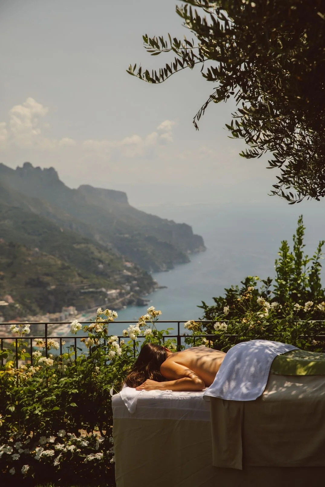 A woman lying face down on an outdoor massage table, surrounded by white flowers and greenery, overlooking a coastal landscape with cliffs and water under a cloudy sky.