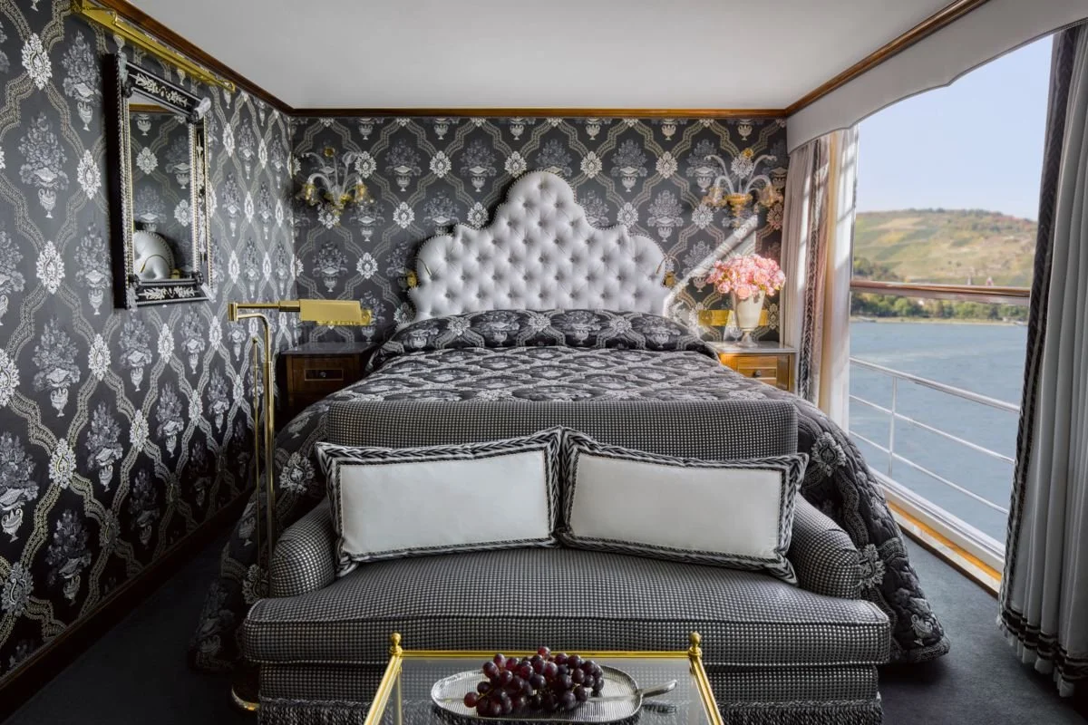 Luxurious bedroom with a black and white wallpapered wall, white tufted headboard, and large window overlooking a river and hills. The room features black patterned bedding, a small sofa with white pillows, a glass-topped table with grapes, a pink-flowered vase, and gold accents.