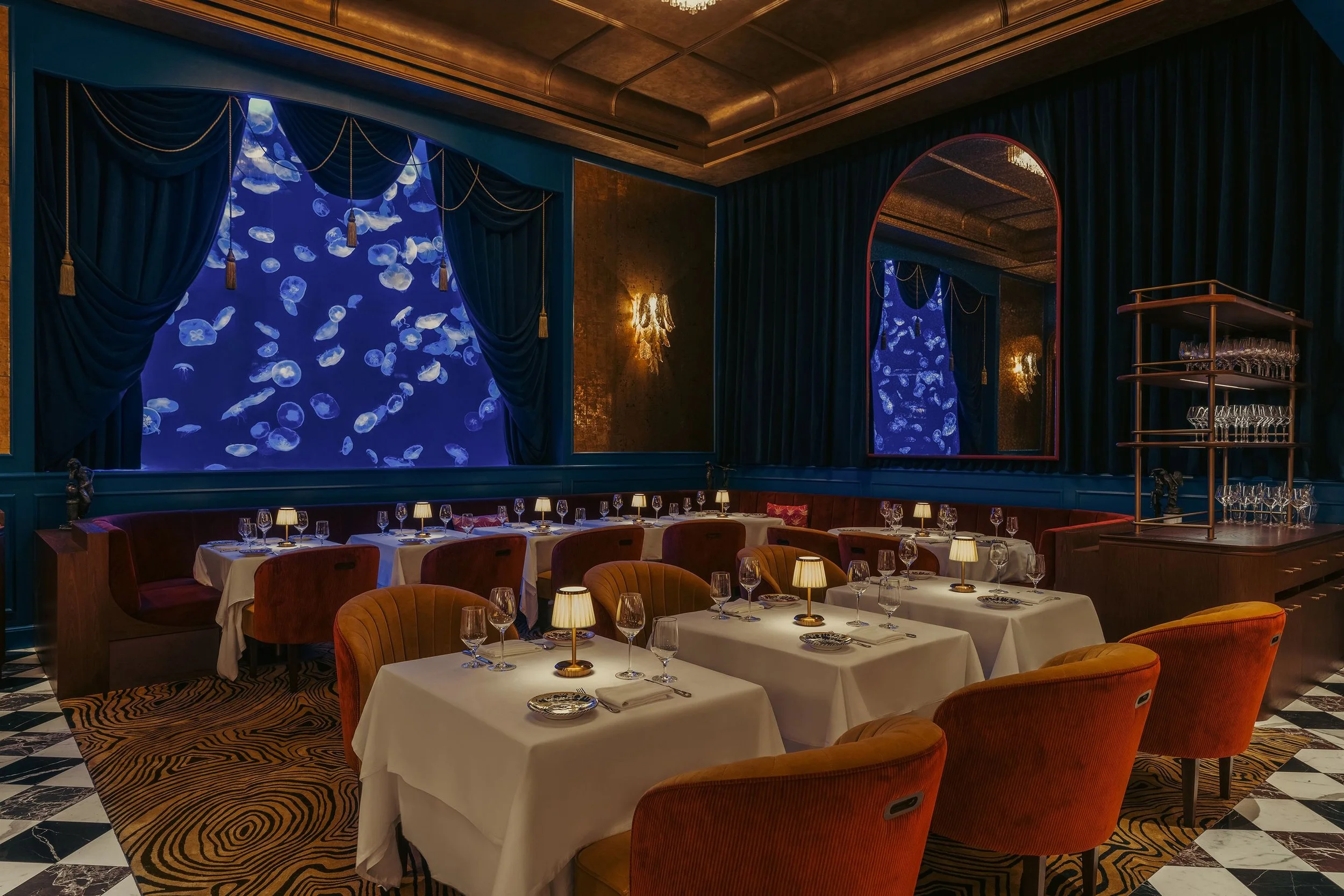 Elegant restaurant interior with tables covered in white tablecloths, set with glasses, plates, and cutlery. Warm lighting, velvet curtains, and a wall with projections of jellyfish create a luxurious atmosphere.
