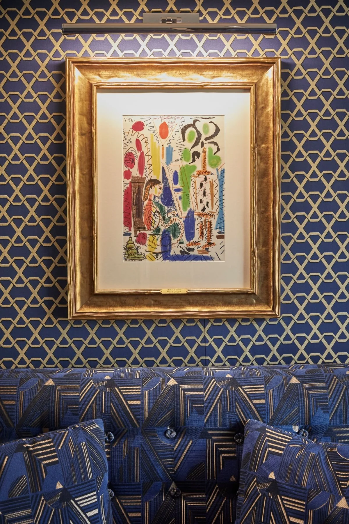 A colorful abstract artwork in a gold frame hanging on a patterned blue and gold wall, with a blue geometric-patterned sofa underneath.
