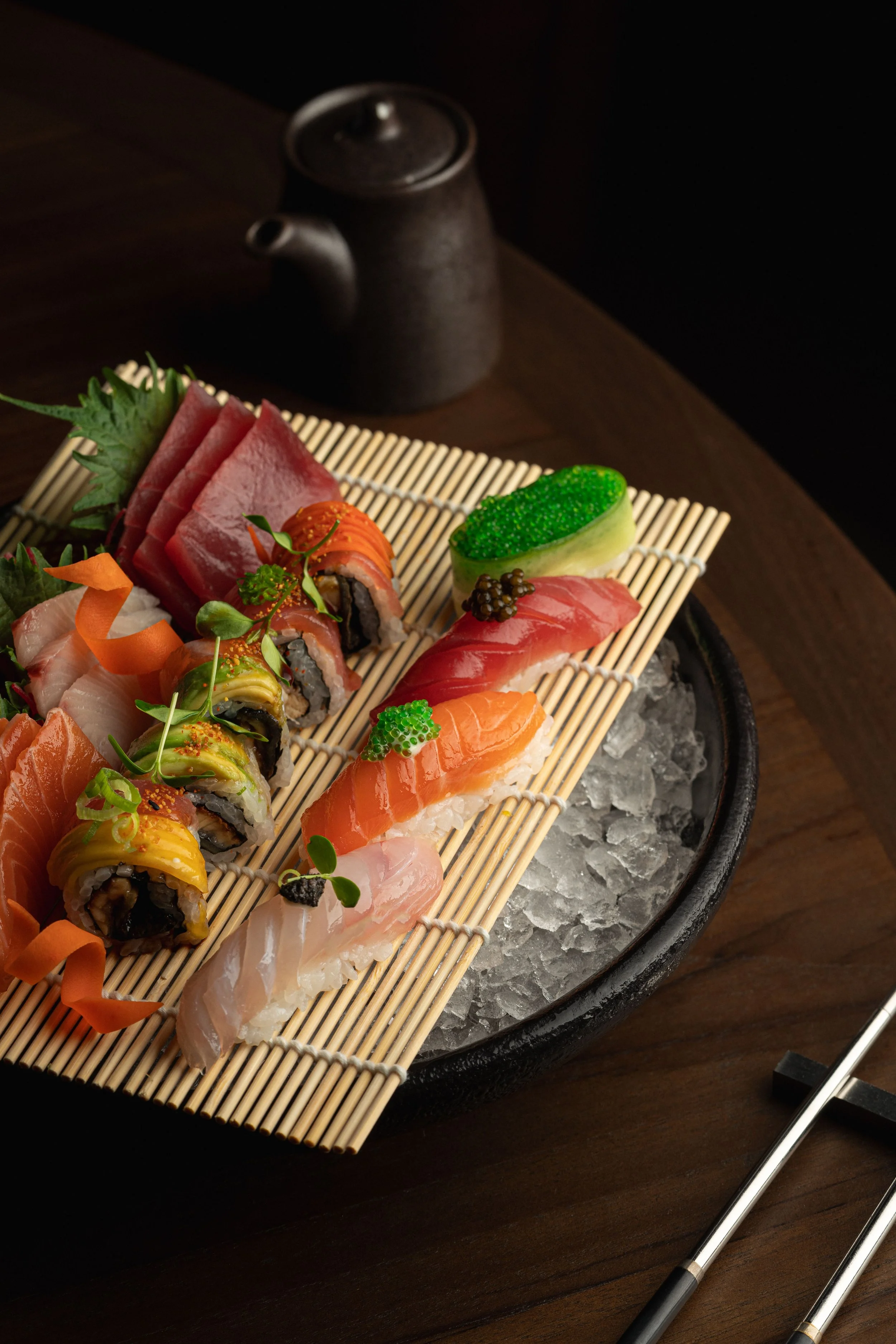 A sushi platter with various types of sushi and sashimi, garnished with wasabi, ginger, and fish roe, placed on a bed of crushed ice, with black soy sauce teapot and chopsticks nearby.