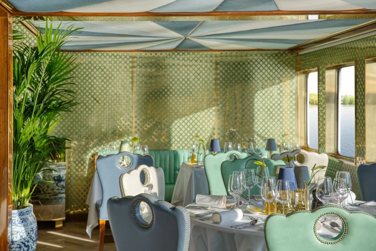 Elegant dining area with pastel-colored chairs, set tables with glassware, cutlery, and blue lamps, surrounded by large windows and greenery outside.