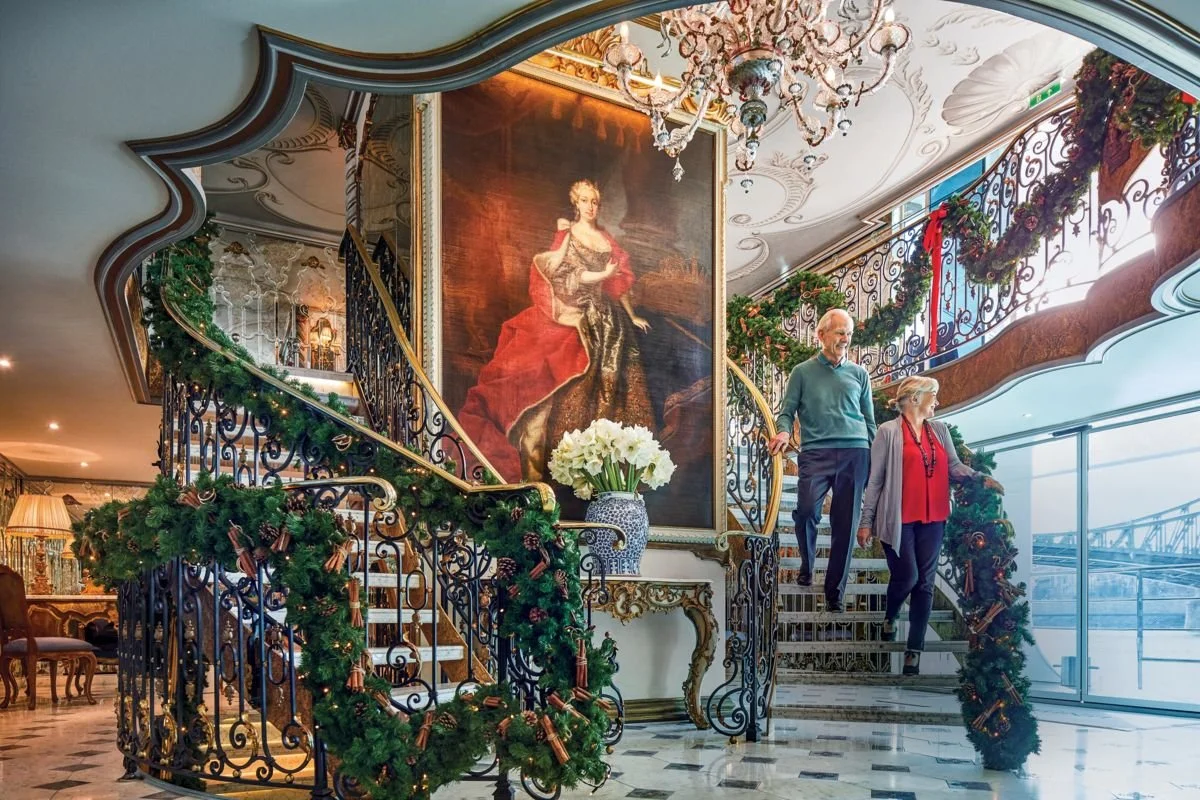 An elegant interior with a spiral staircase decorated for Christmas, featuring green garlands with ornaments, and a large classical portrait of a woman in regal attire displayed on the wall. An elderly man and woman walk down the staircase, holding hands.