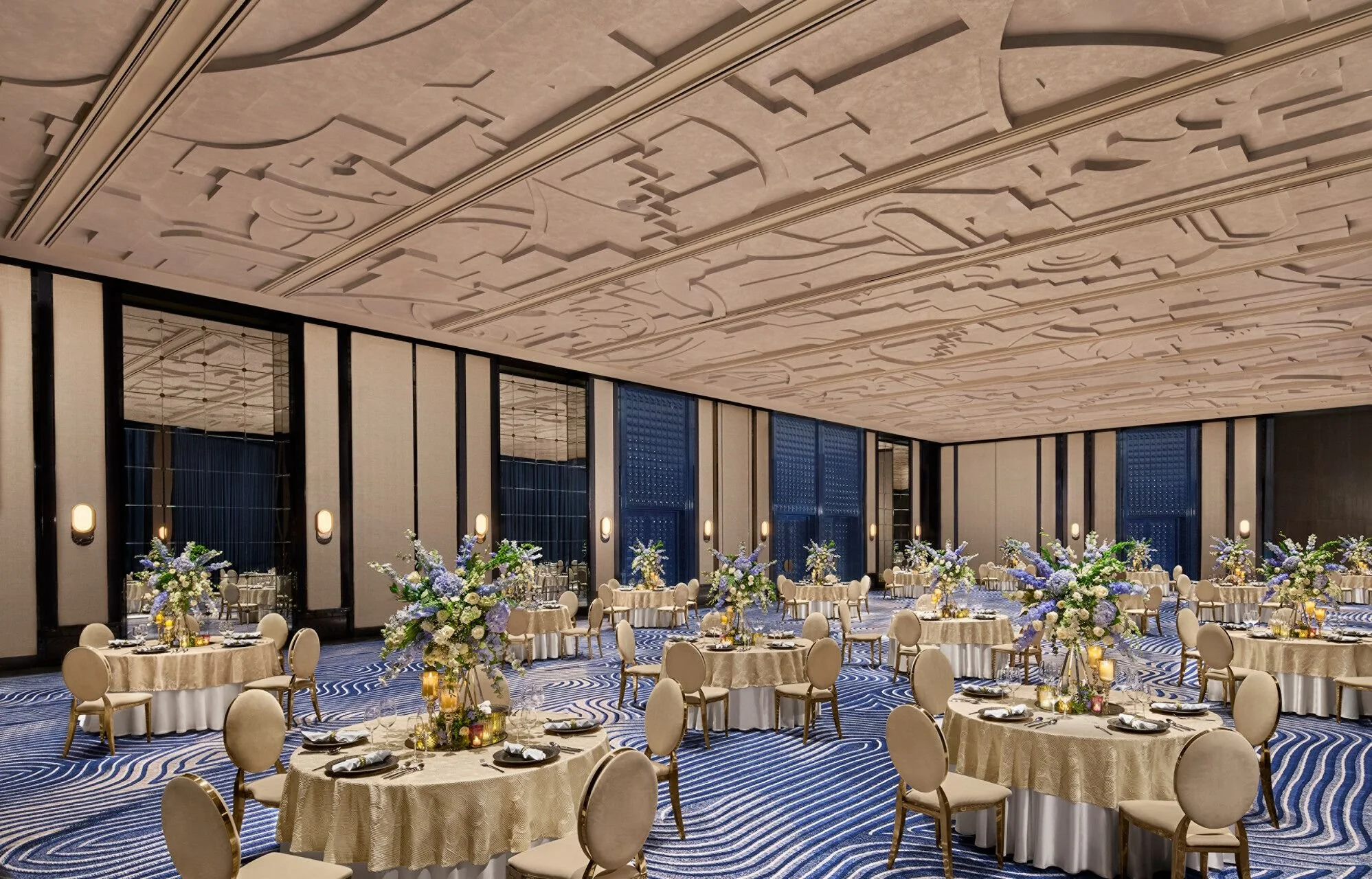 Elegant banquet hall decorated with floral centerpieces, round tables, beige chairs, and a high ceiling with geometric designs.
