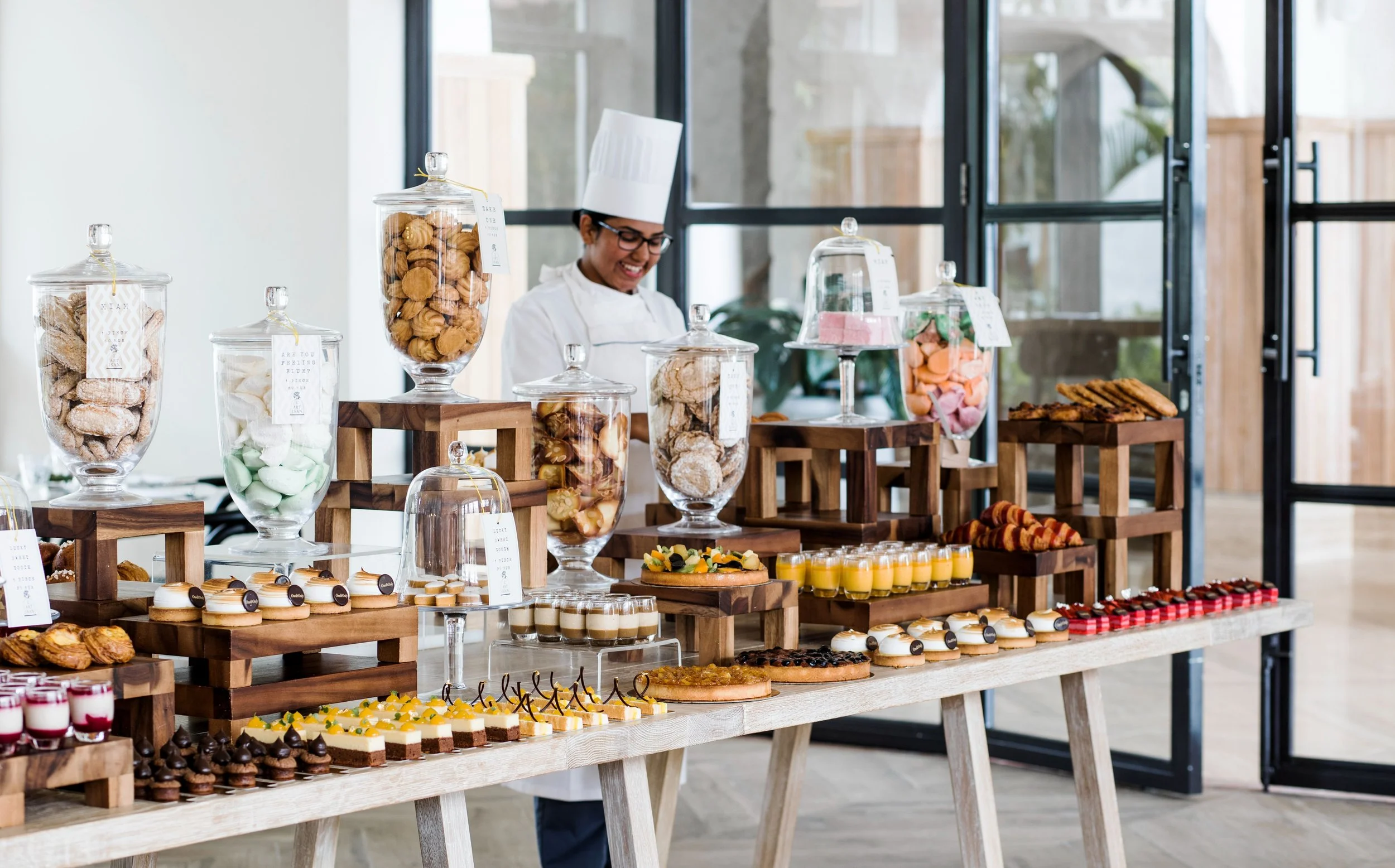 A woman wearing a chef's hat and glasses is smiling behind a display of various cookies, pastries, and desserts arranged on wooden stands with glass jars containing sweets. The setting appears to be a modern, well-lit indoor space with large windows.