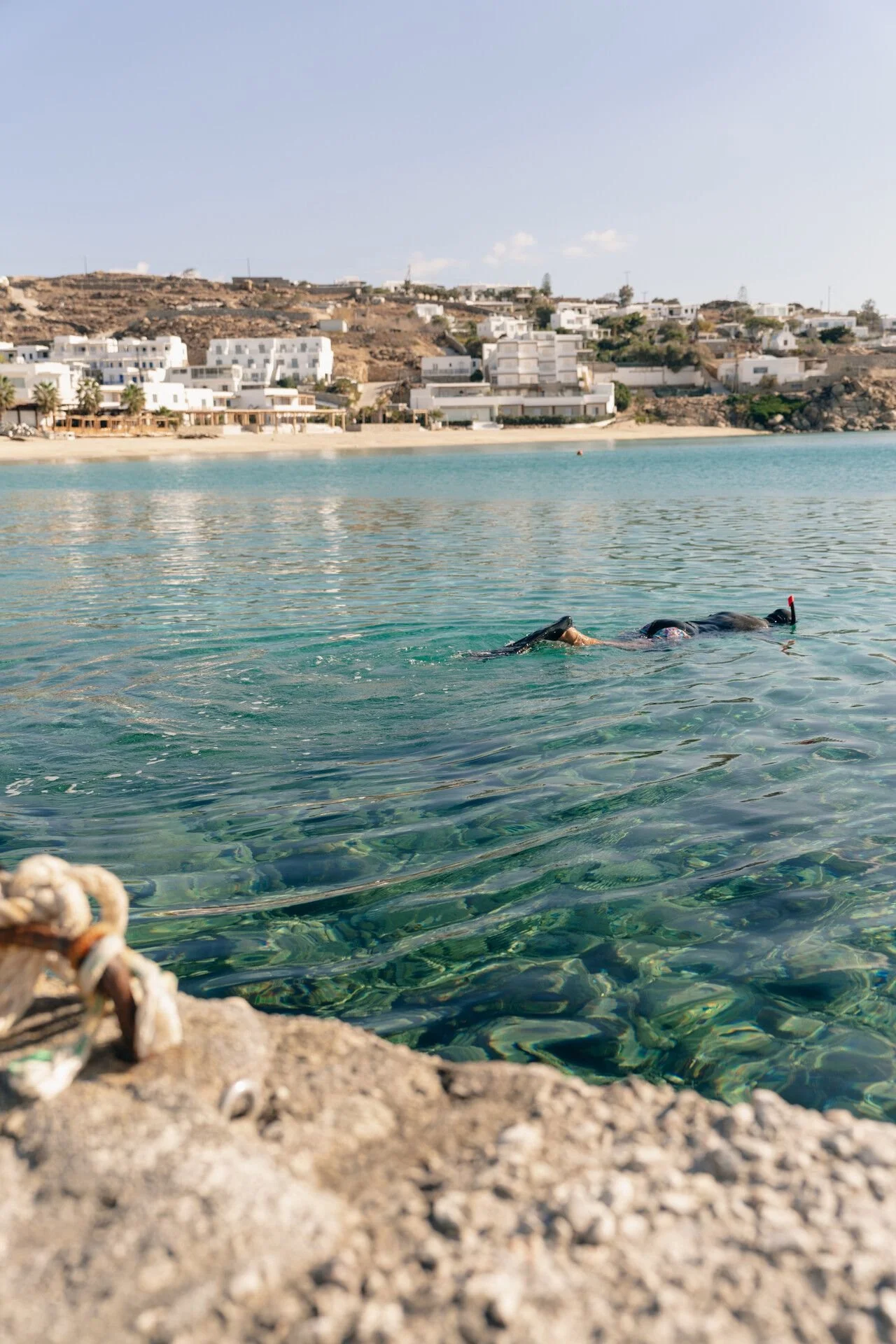 Person snorkeling in clear water near rocky shoreline with white buildings and hills in the background.