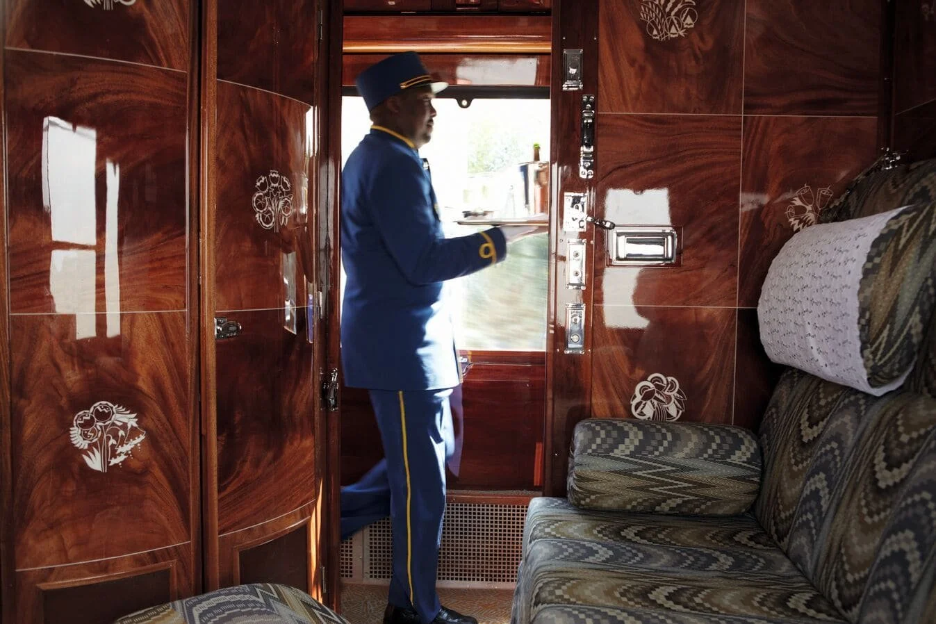 A train conductor in a blue uniform and hat standing near the door of a vintage train car with wood-paneled walls, looking outside through the open door.