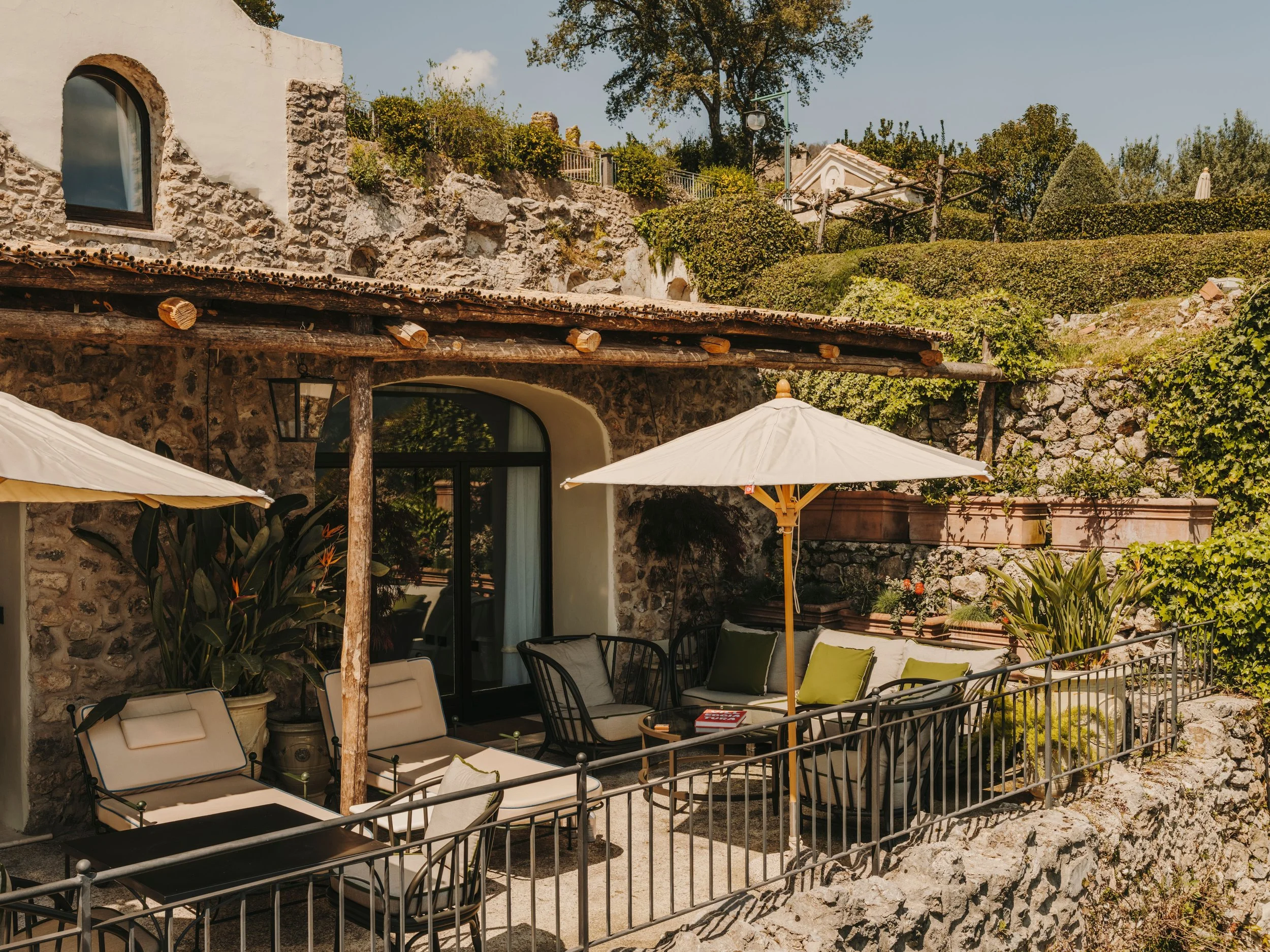 Outdoor patio area with lounge chairs, table with a book, large yellow patio umbrella, lush green plants, a stone and stucco building with large arched window, and greenery on the hillside in the background.