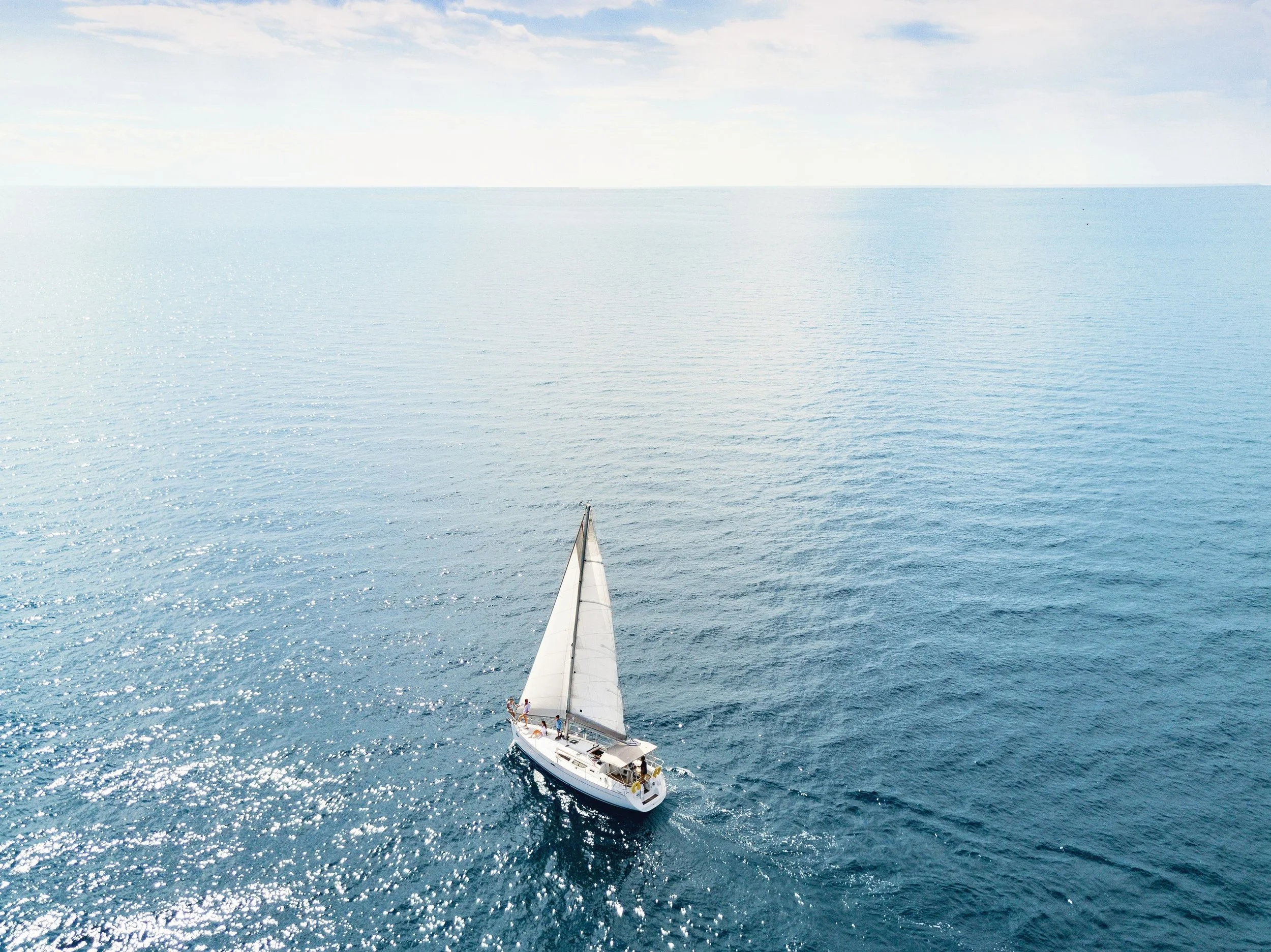 A sailboat with people on it sailing in the open ocean under a partly cloudy sky.