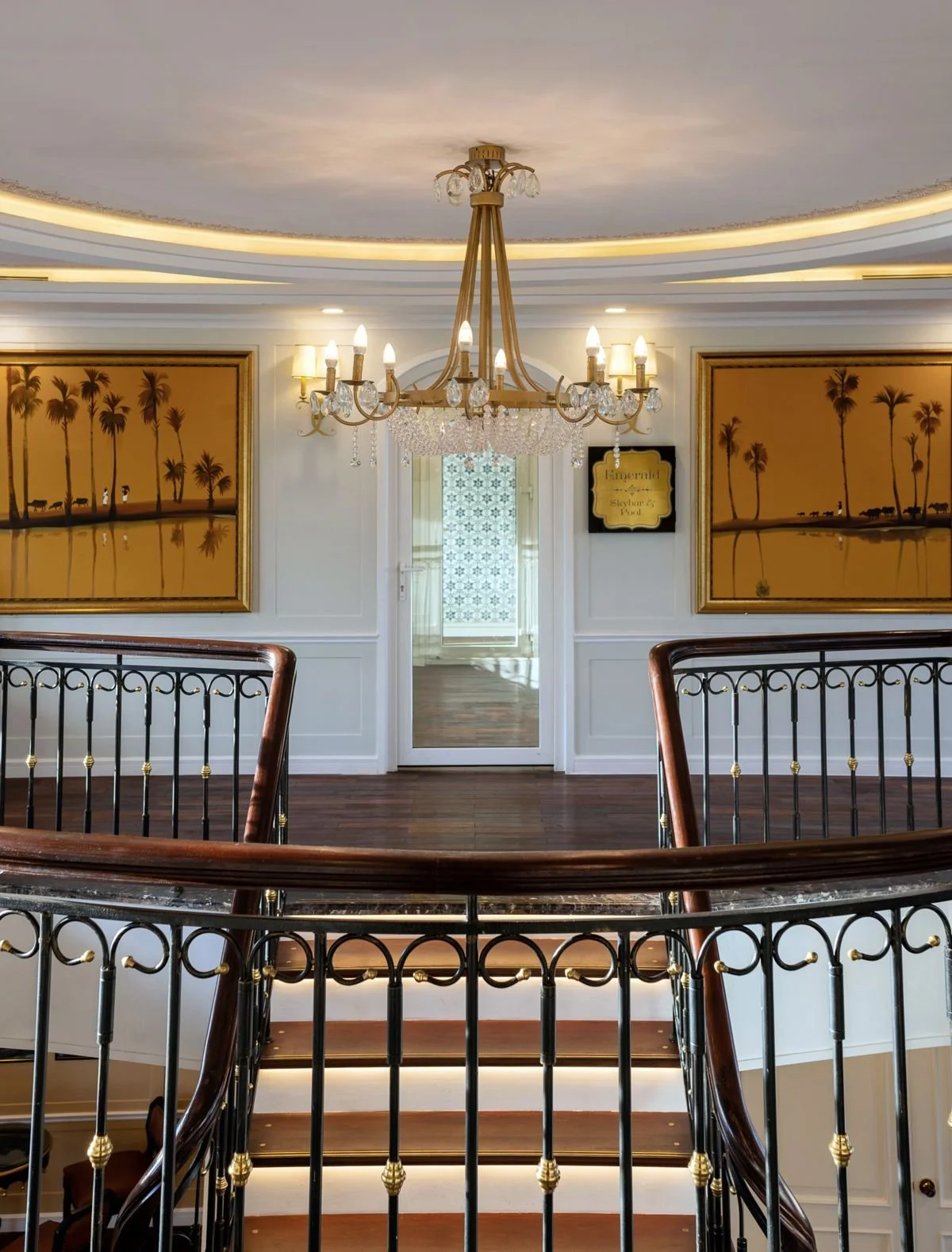 Interior view of a staircase landing with a chandelier hanging from a decorated ceiling. There are two framed paintings with palm trees on the wall and a mirrored door leading to another room with patterned windows.