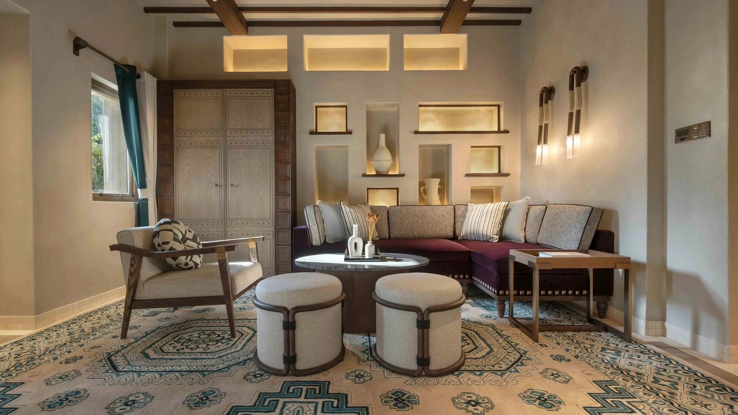 Living room with a large sectional sofa, a round coffee table with decorative vases, a single armchair, and built-in wall niches with decor. The room has wood-beamed ceiling, patterned rug, and a window with teal curtains.