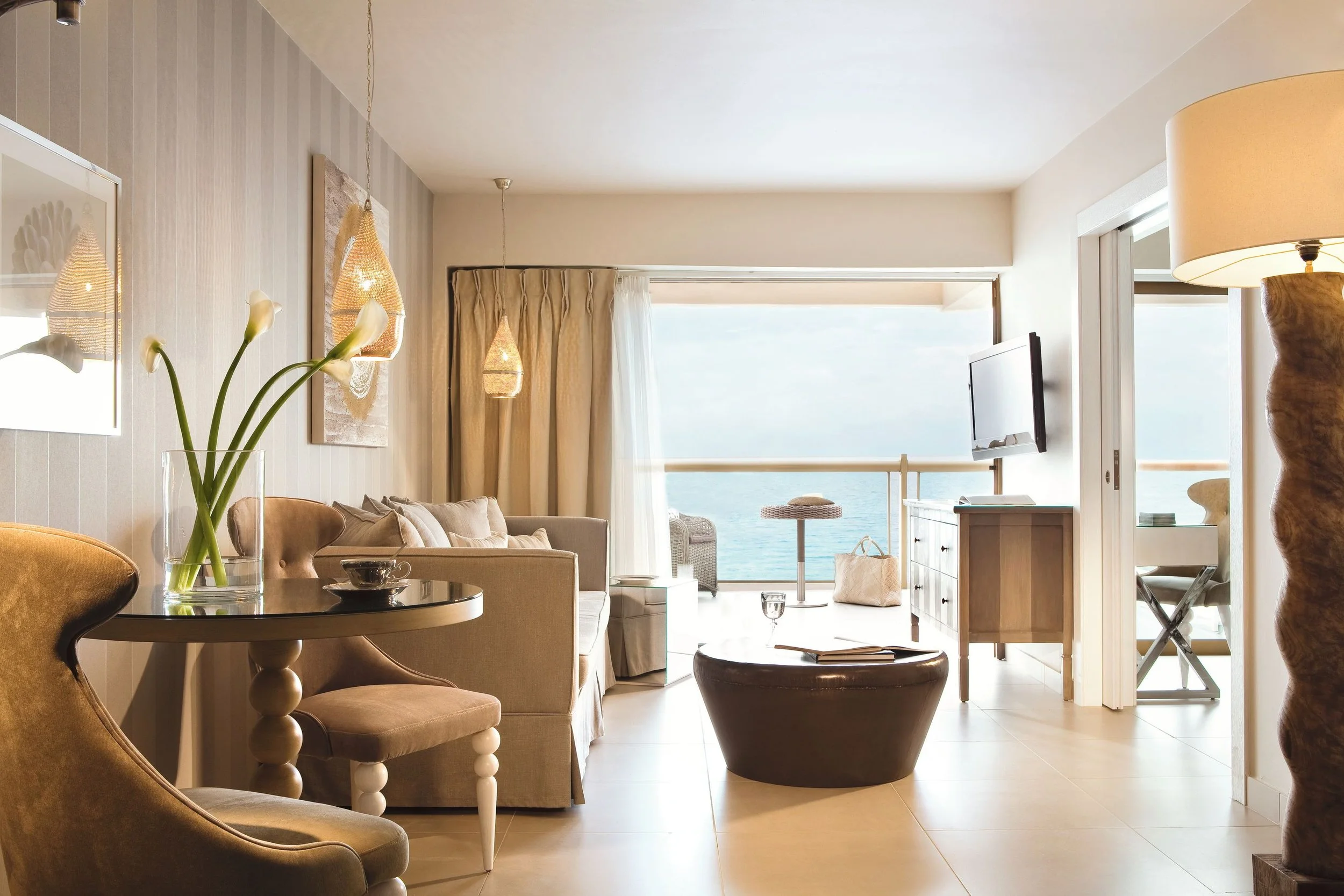 A living room with light-colored curtains, a beige sofa, a round black coffee table, and a glass vase with white flowers on a dining table. There are hanging woven pendant lights, a television, a large window with a view of the ocean, and a door leading to a balcony with outdoor furniture.