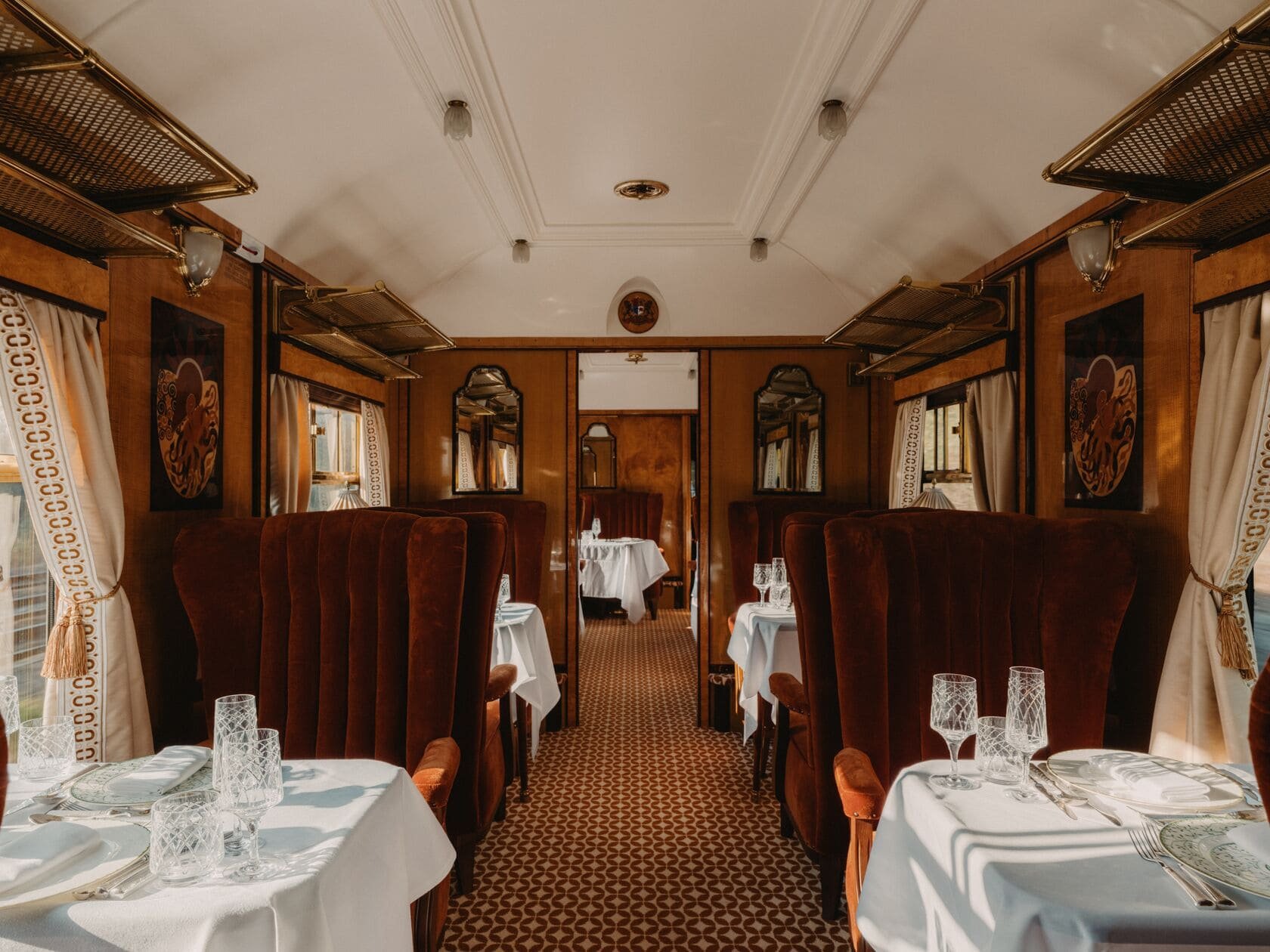 Interior of a vintage train dining car with white tablecloths, fine glassware, and ornate curtains, featuring wood-paneled walls, mirrors, and upholstered chairs.