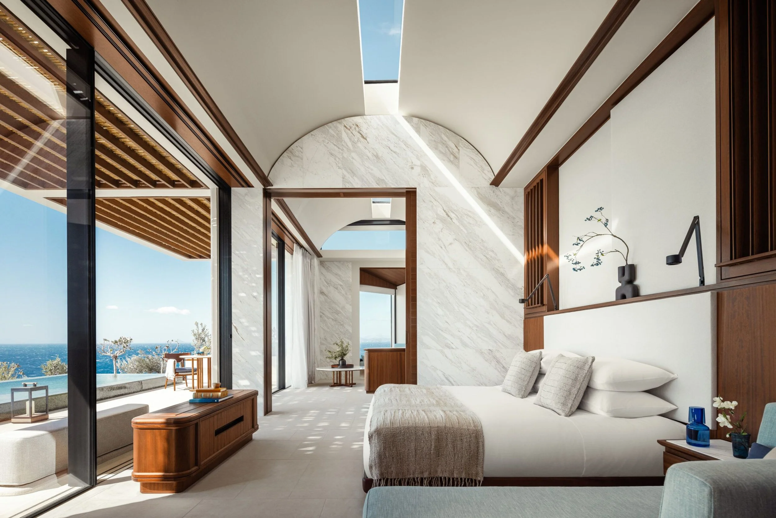 Modern living room with large glass sliding doors opening to an outdoor patio with ocean view. The room features a white sofa, a wooden coffee table, and elegant decor including a vase with flowers and wall art.