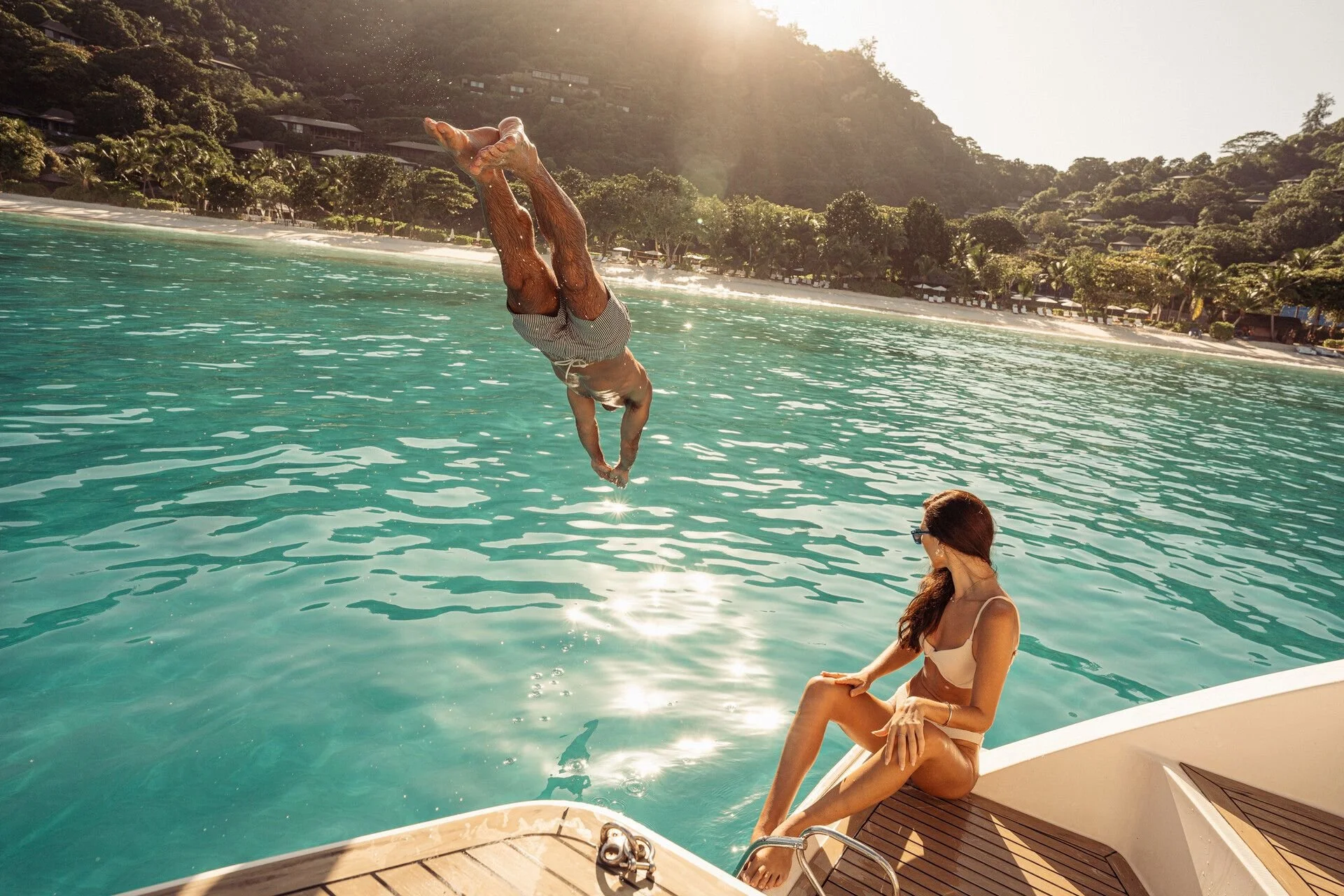 A person jumping into turquoise water from a boat with another woman sitting on the boat, while a scenic beach with trees and hillside houses is visible in the background, sun shining brightly.