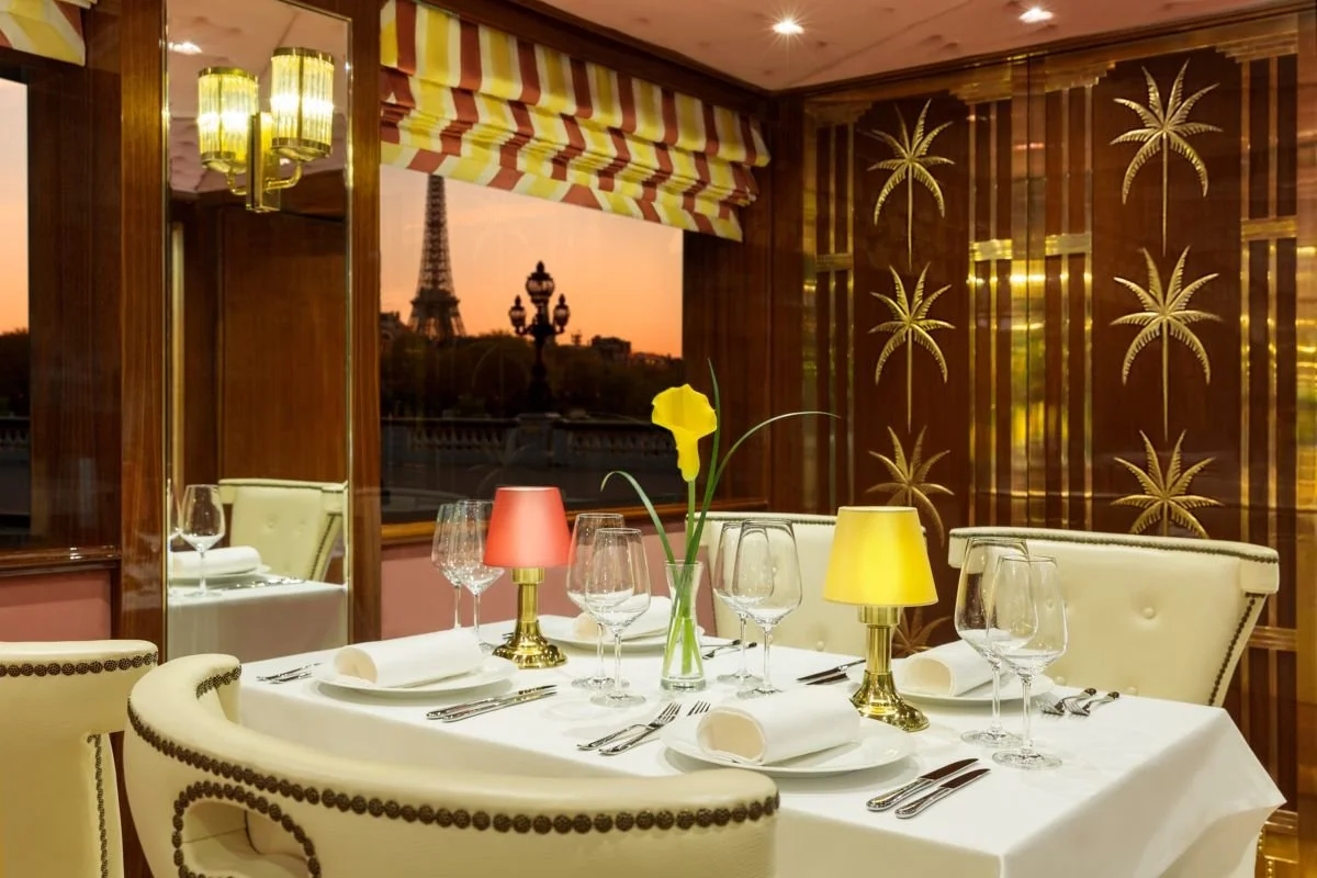 Elegant dining room with a table set for four, featuring white tablecloth, glassware, silverware, napkins, and small lamps with yellow and pink shades, a single yellow flower in a vase, and a window overlooking Paris with the Eiffel Tower at sunset, decorated with palm tree gold patterns on the wall.