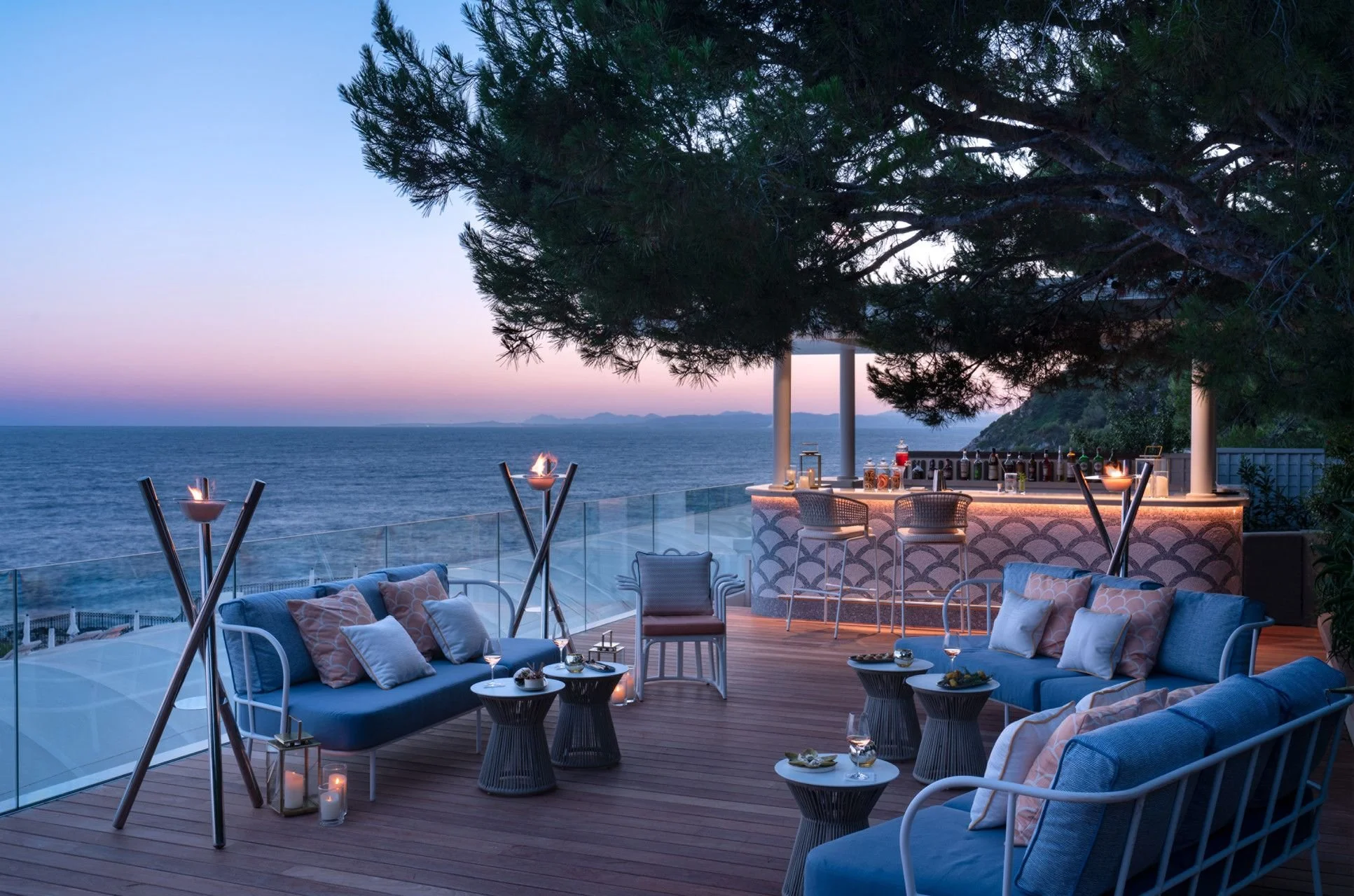 Luxury outdoor bar area on a deck overlooking the ocean at sunset, with comfortable seating, tiki torches, and a large tree providing shade.