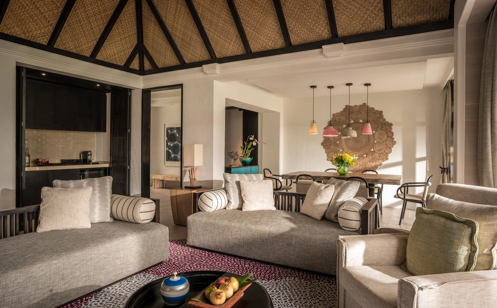 A spacious living room with beige sofas, a round wooden table with an apple centerpiece, and abstract wall art. The room features a high ceiling with wooden beams, a dining area with a wooden table and black chairs, and pendant lights hanging from the ceiling.