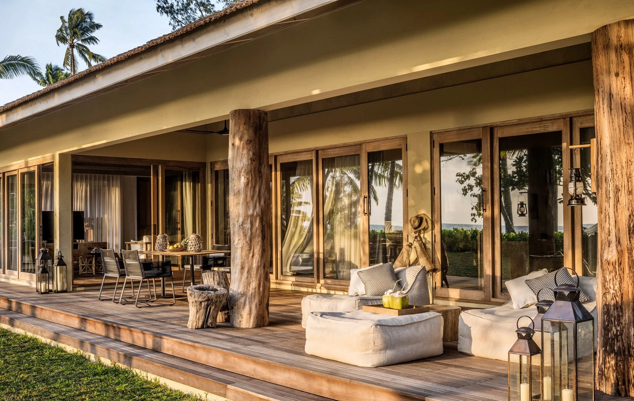A wooden house with large glass doors and a deck, surrounded by palm trees, with outdoor furniture including chairs, sofas, and lanterns, and the ocean visible through the glass doors.