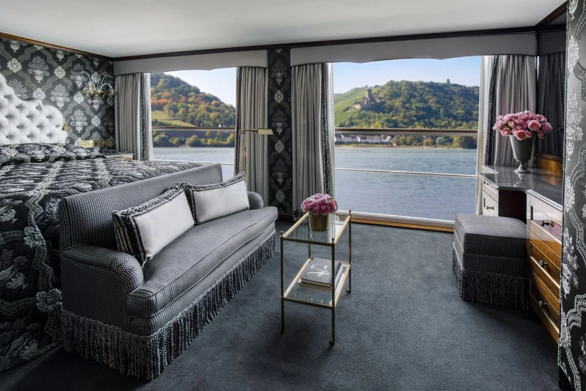 Luxurious hotel room with large window view of a river and green hills, black and white patterned wallpaper, a bed with black and white bedding, a checkered sofa with throw pillows, a glass-topped side table with pink flowers, and a dresser with a pink flower arrangement.