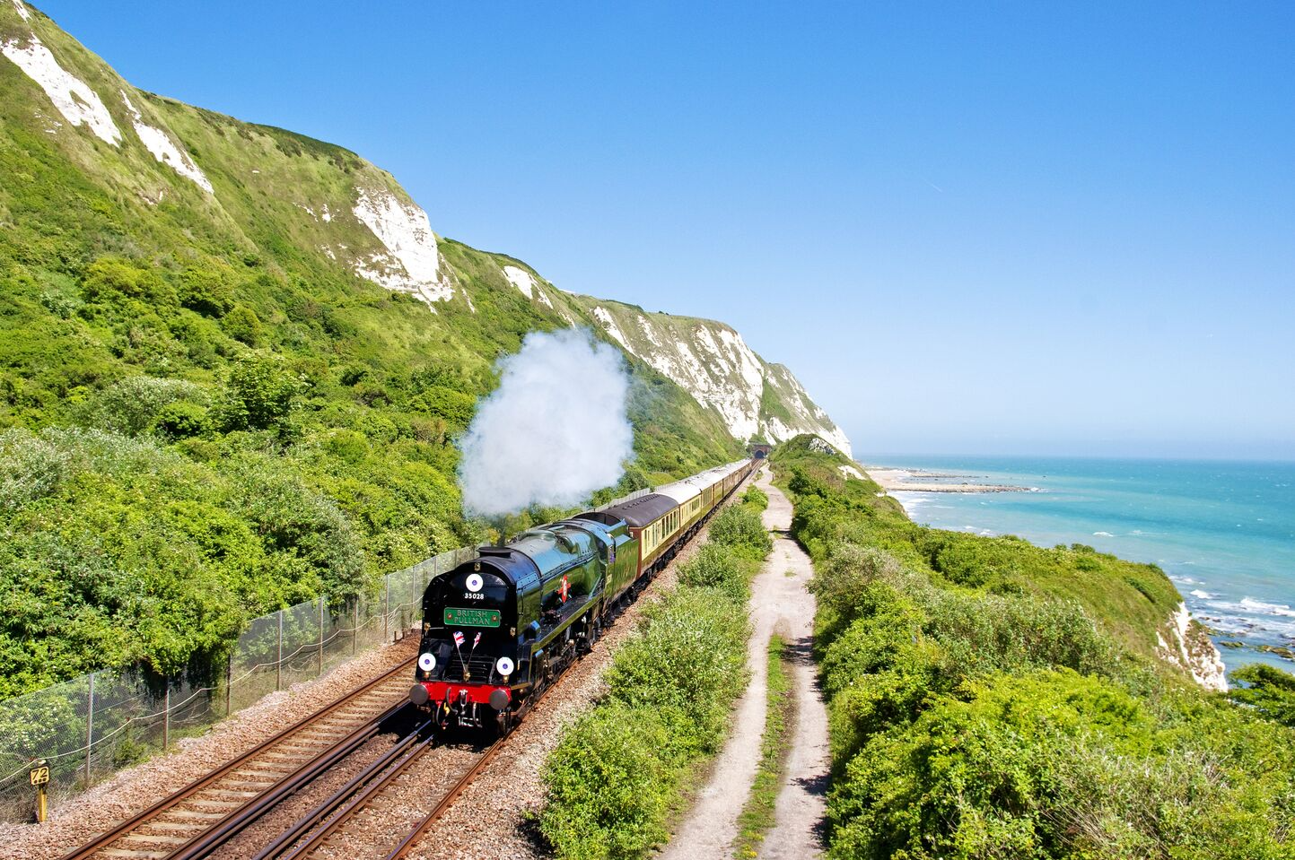 A vintage steam train traveling along the coast with green cliffs on one side and the ocean on the other, under a clear blue sky.