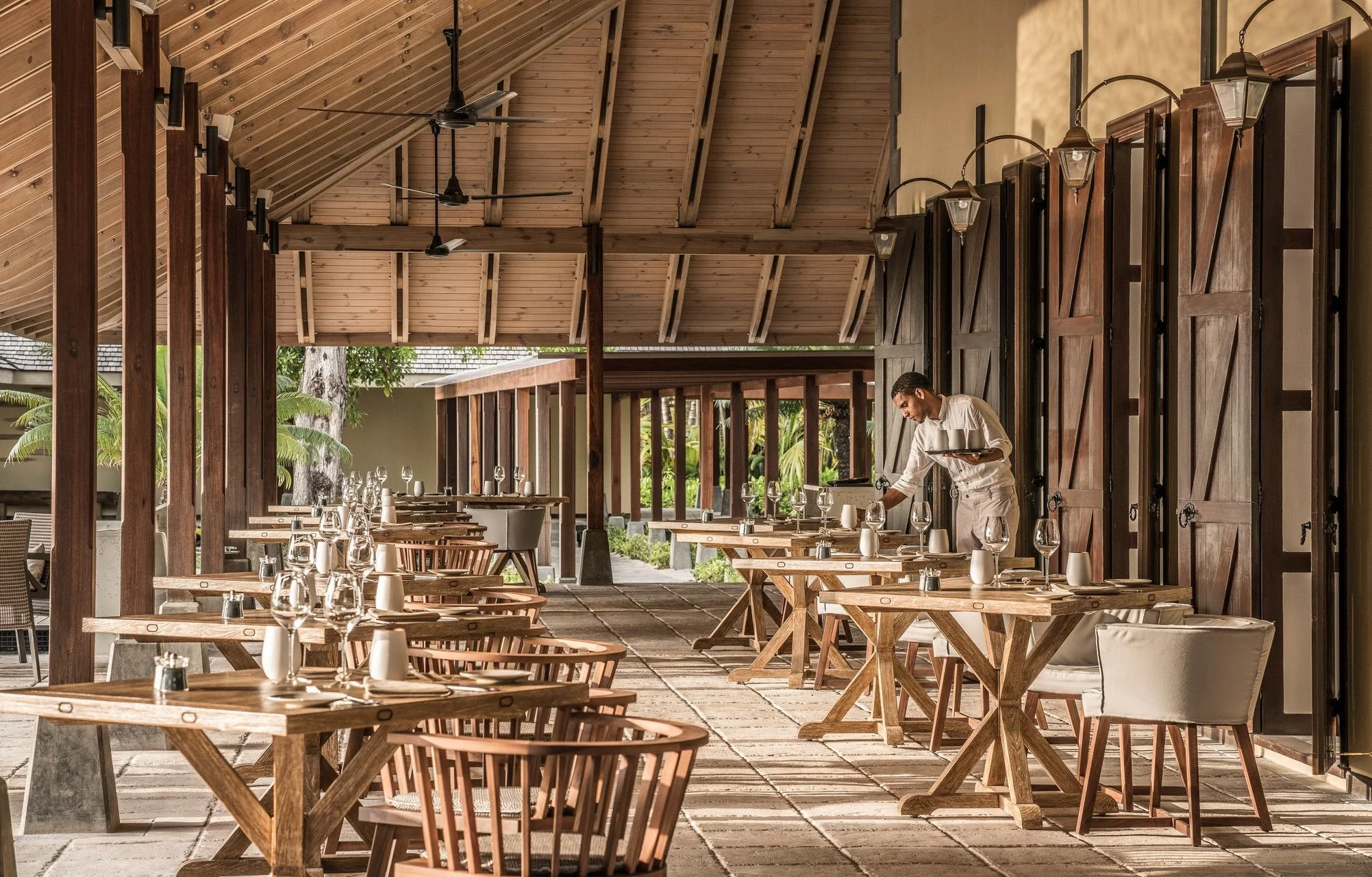 Empty outdoor restaurant dining area with wooden tables and chairs, a waiter setting tables, and a wooden ceiling with fans and hanging lanterns.