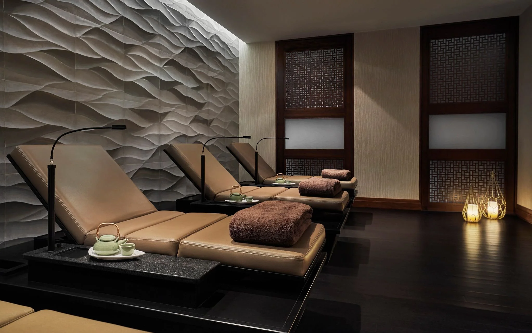 A relaxing spa room with three beige lounge chairs, each with a matching towel, small tray with teapot and cups, black lamps overhead, decorative textured wall, wooden sliding doors with intricate lattice design, and glowing lanterns on the dark wooden floor.
