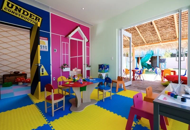 Colorful indoor children's playroom with various toys and furniture, open door leading to an outdoor play area with a slide and shaded roof.