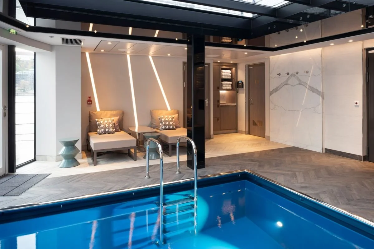 Indoor pool area with lounge chairs, a small side table, and a wall with modern lighting and decorative panels.