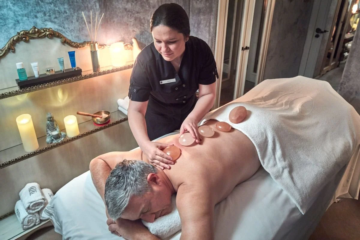 A woman is giving a hot stone massage to a man lying face down on a massage table in a spa room, with black stones placed along his back and a therapist applying more stones.