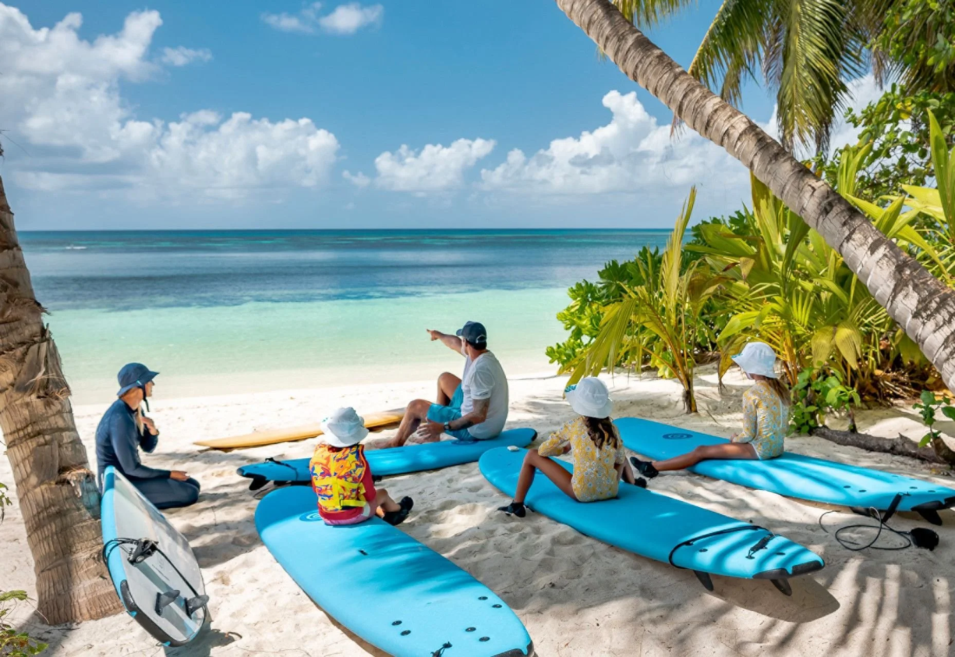 A group of people on a sandy beach with palm trees, some on surfboards, and the ocean in the background. One man is sitting on a blue surfboard, pointing towards the ocean, while others sit or stand nearby, some wearing hats and colorful clothing.