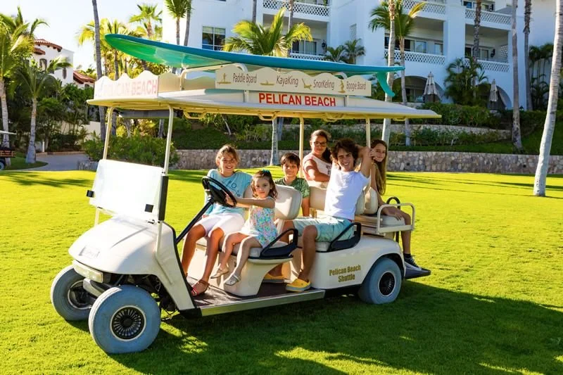 Six people sitting on a golf cart with a canopy that reads "Pelican Beach" and has a green paddle board on top, parked on a grassy area near tall palm trees and a white building.