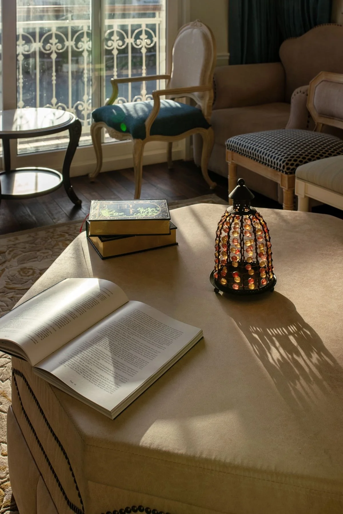 Sunlight streams into a cozy living room with a table holding a decorative lantern, an open book, and a stack of books. In the background, there are chairs and a sofa near a balcony with ornate railing, creating a warm and inviting atmosphere.