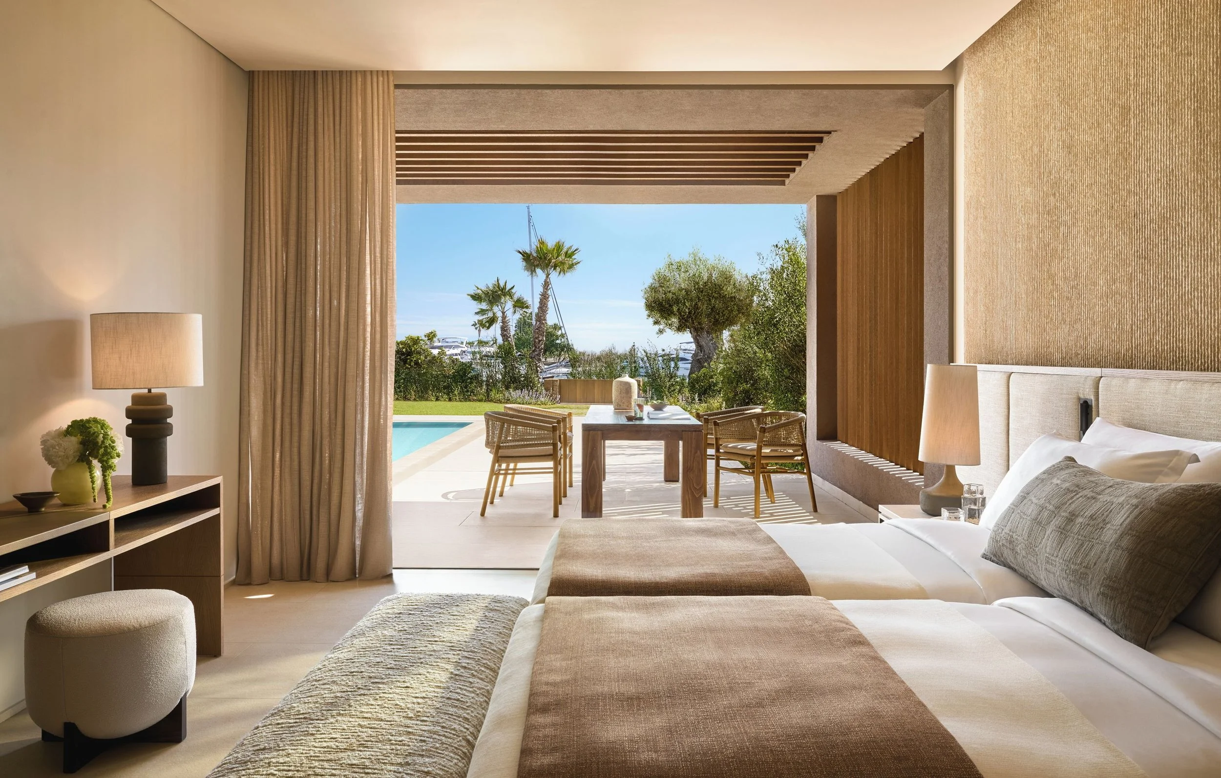A hotel room with a king-sized bed, sitting area, and an open wall leading to a patio with a view of a garden, pool, and palm trees.