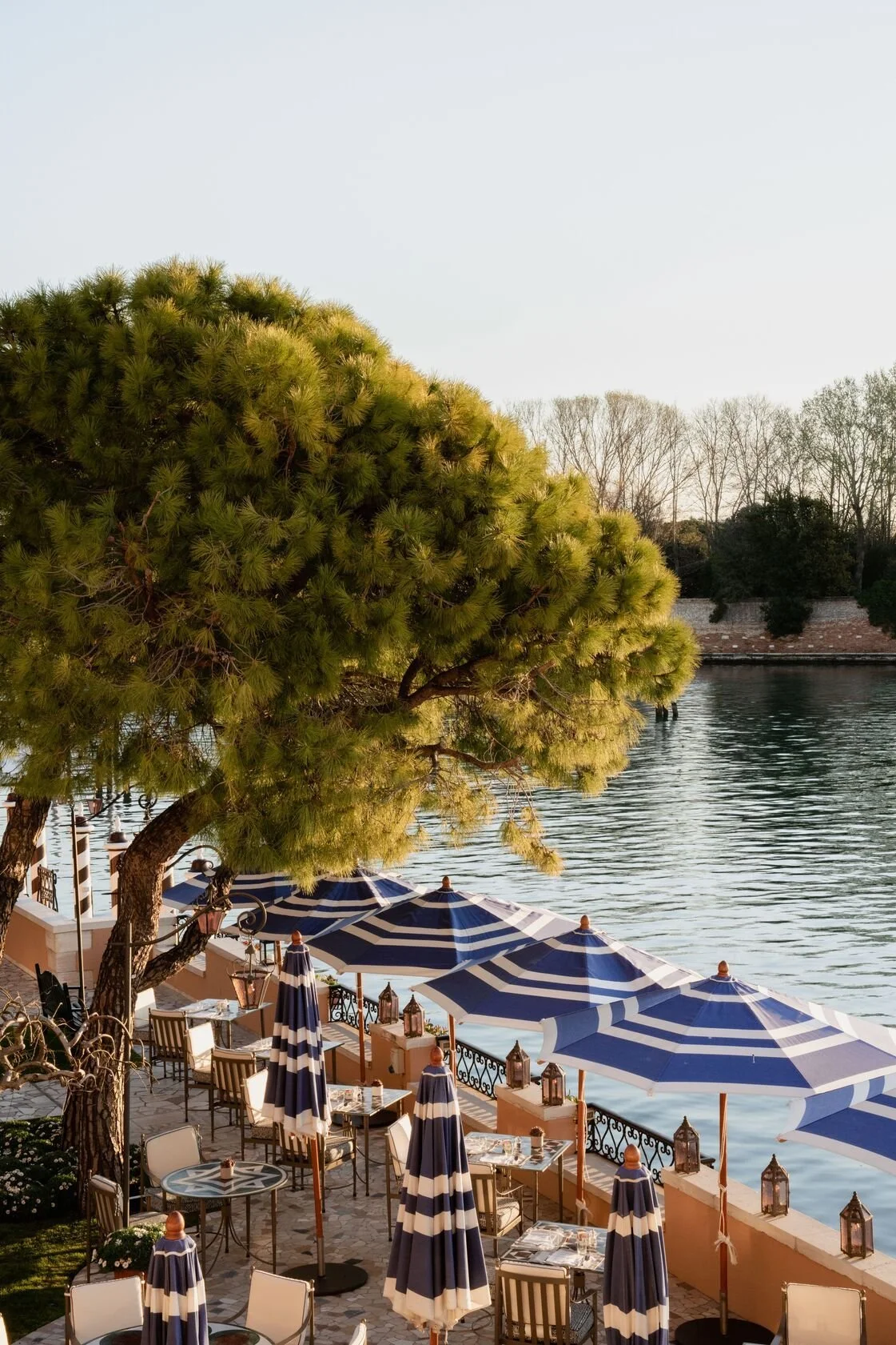 Outdoor dining area with blue and white striped umbrellas, tables, and chairs along a river, with a large tree in the foreground and trees in the background.