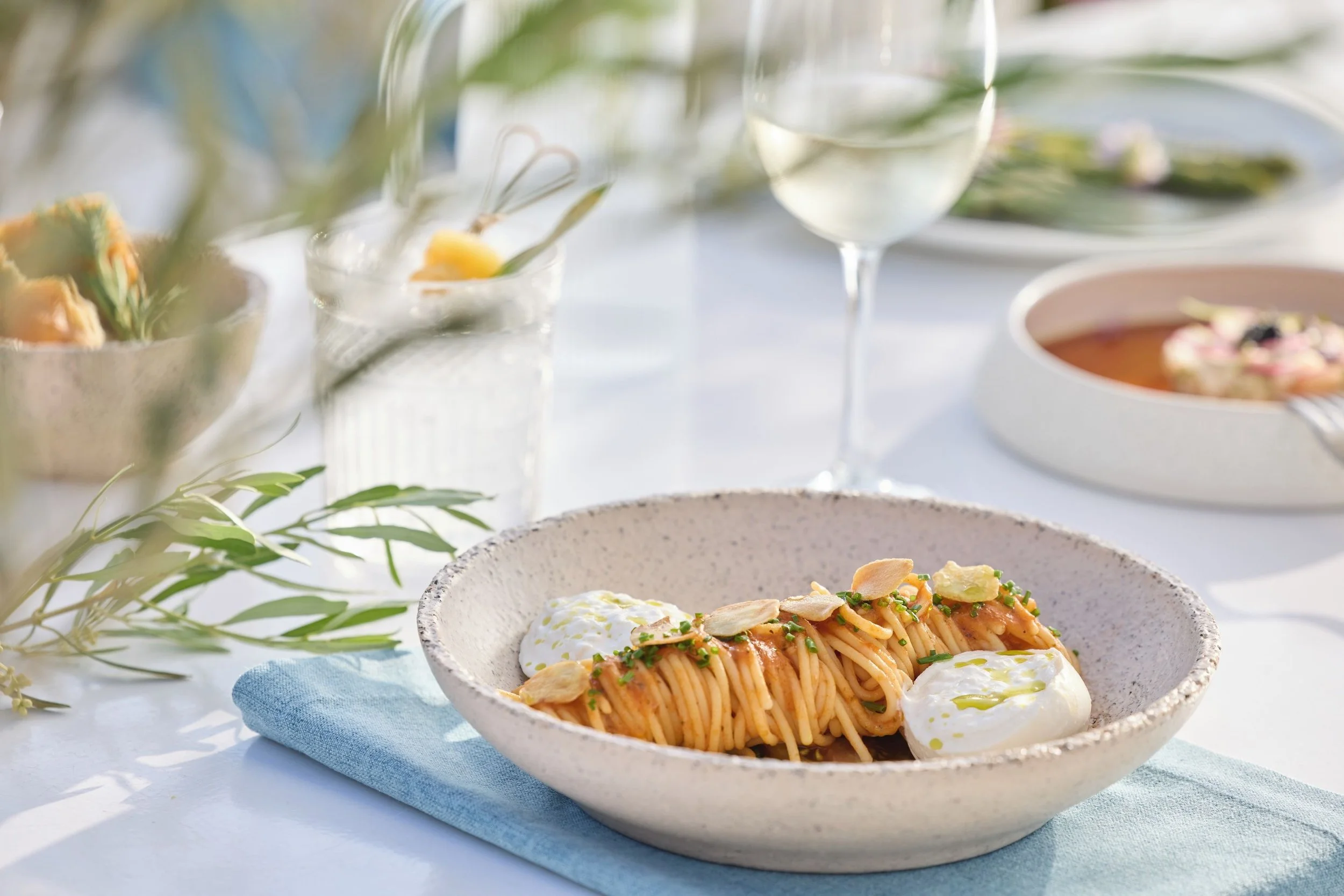 A dish of spaghetti with tomato sauce, topped with sliced almonds and garnished with herbs, served with dollops of cream or cheese on a speckled ceramic bowl on a blue napkin.
