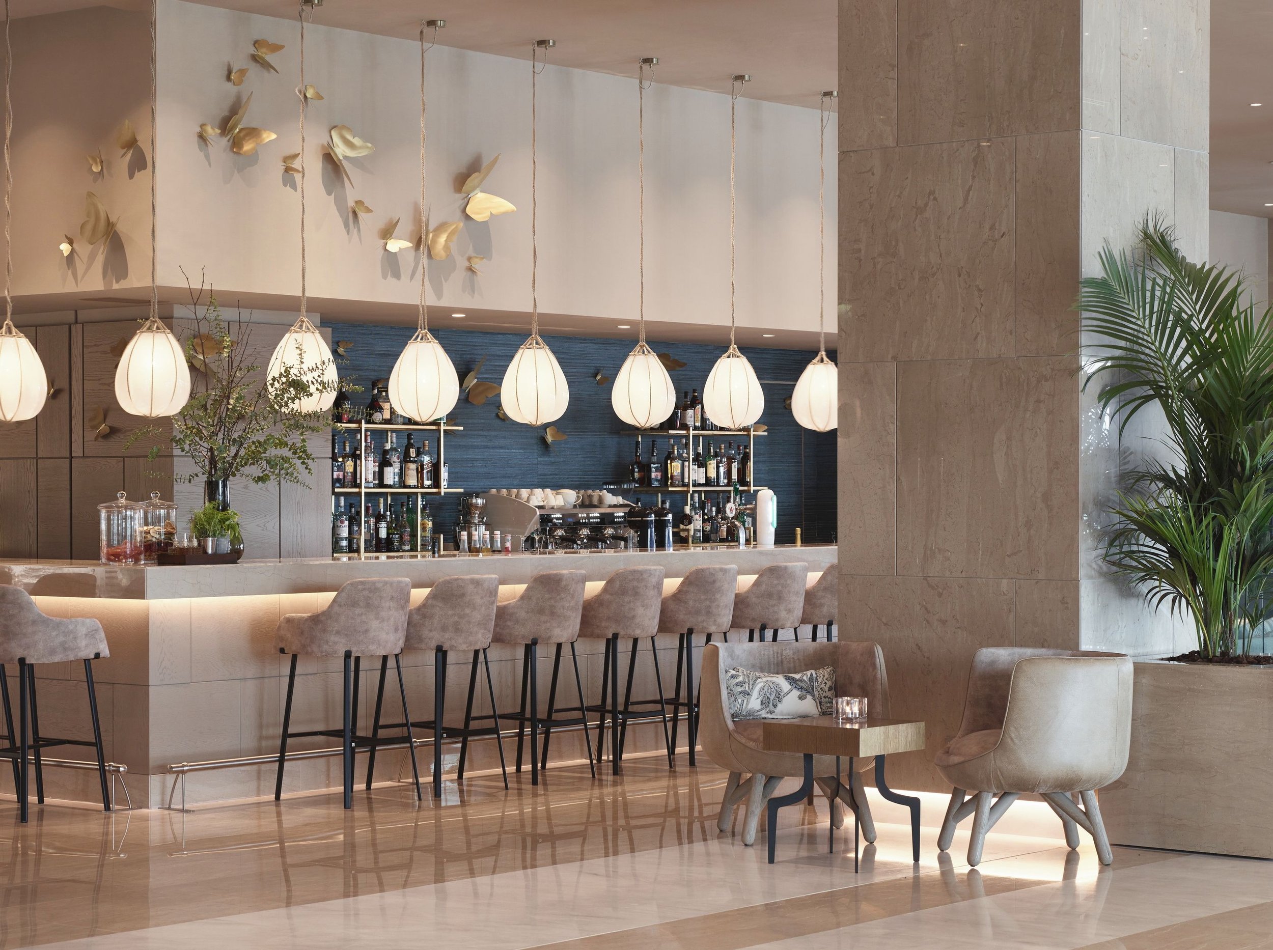Interior of a modern restaurant or bar with a marble bar counter, beige chairs, hanging pendant lights, a blue accent wall, shelves with liquor bottles, and decorative gold butterfly wall art. There is a large stone column, greenery, and a cozy seating area with a cushion and a glass on a small wooden table.