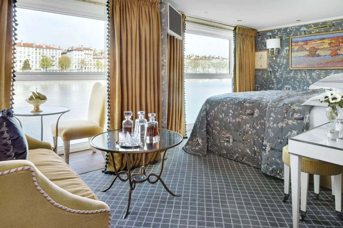 A hotel room with large windows overlooking a river and cityscape, featuring a bed with floral bedding, a white desk with a small vase of flowers, a seating area with a yellow armchair and a small table, and a glass coffee table with bottles and glasses, decorated with curtain drapes and artwork on the walls.