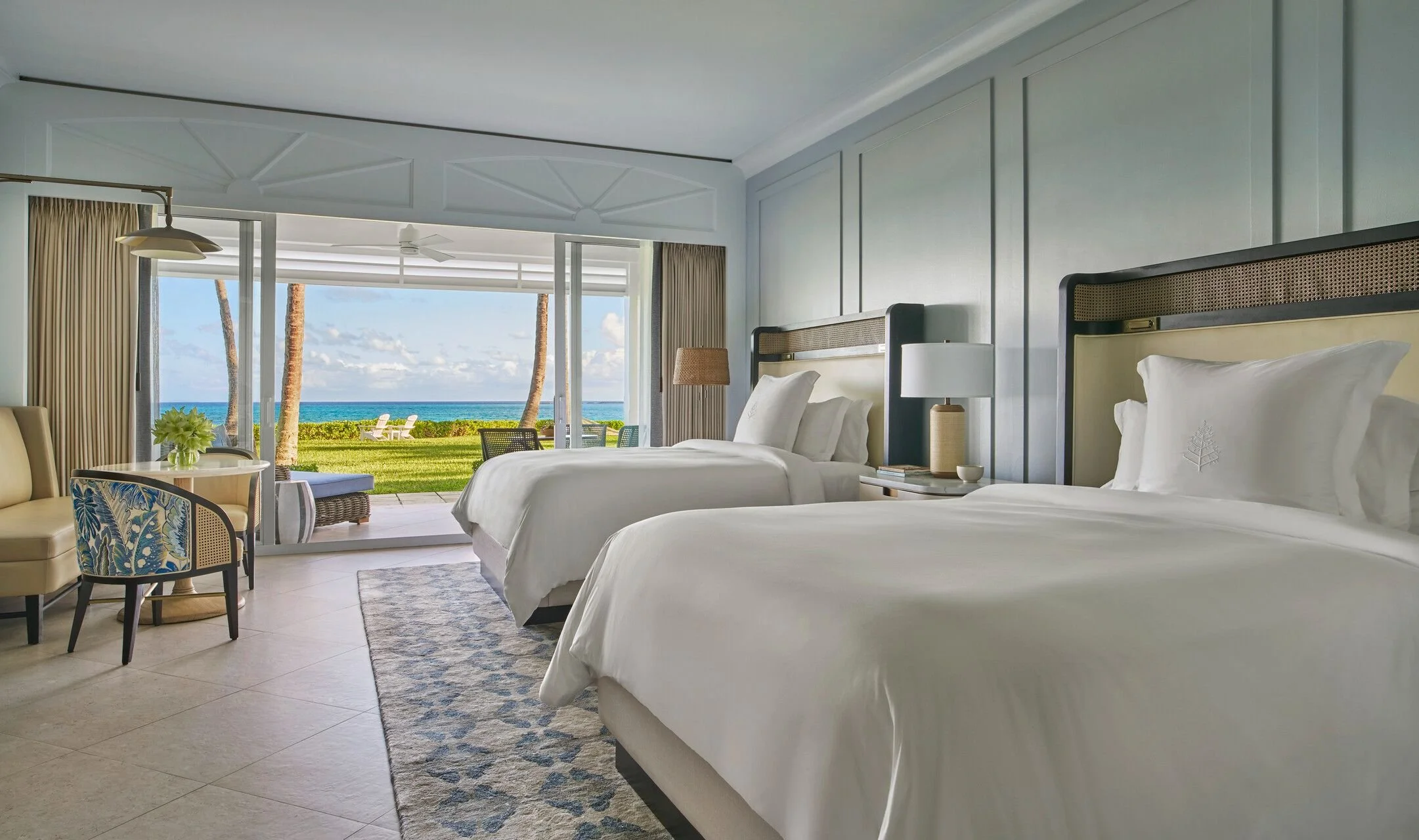 Beach view from a hotel room with two beds, a small sitting area, and sliding glass doors opening to a grassy area with palm trees and ocean.