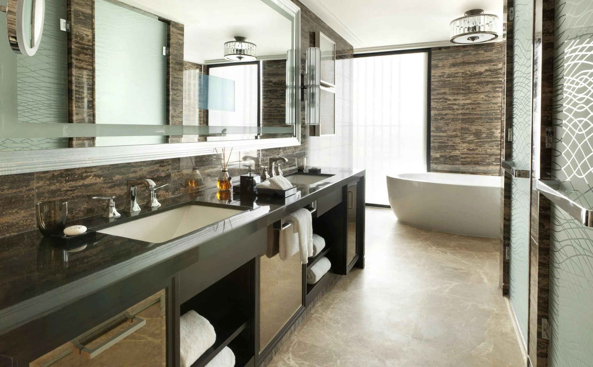 Modern bathroom featuring a large mirror, dual sinks with dark cabinetry, a freestanding bathtub, marble walls, and large windows with blinds.