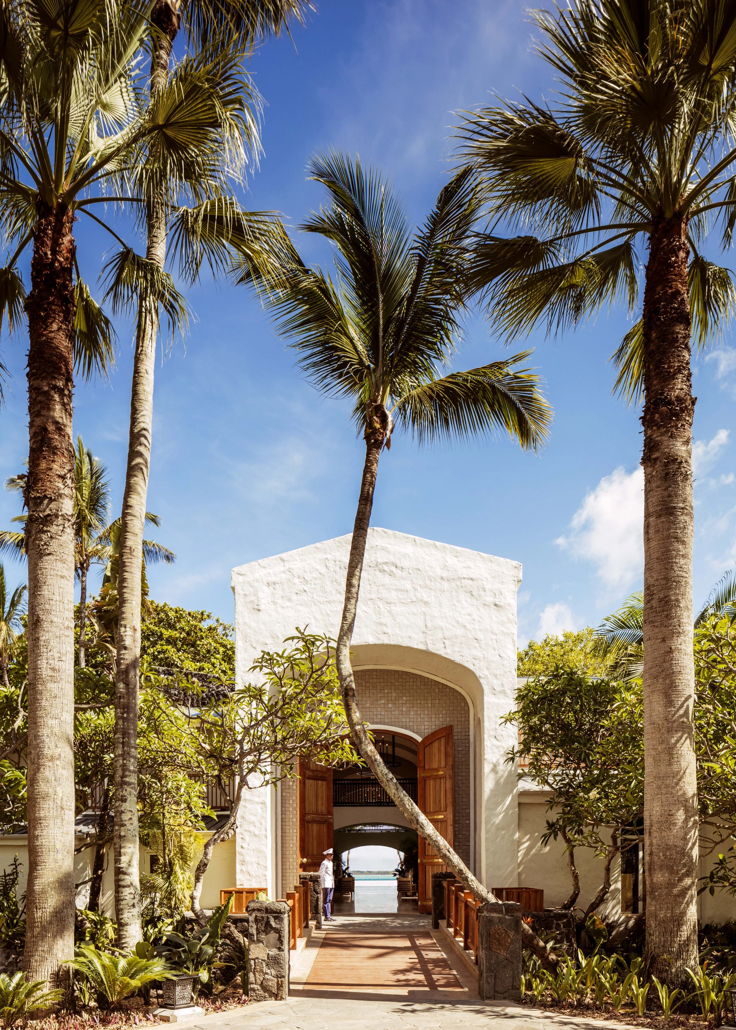 White building with a tiled archway entrance, surrounded by tall palm trees and lush greenery, under a blue sky with scattered clouds.